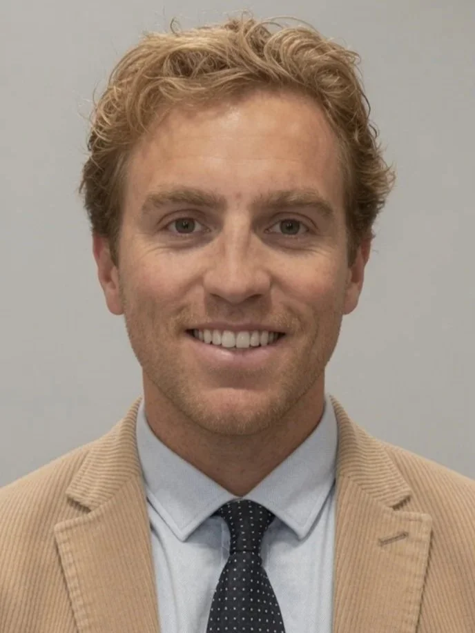 A young man with curly blonde hair, smiling, wearing a beige suit jacket, white shirt, and black polka dot tie, standing against a plain light gray background.