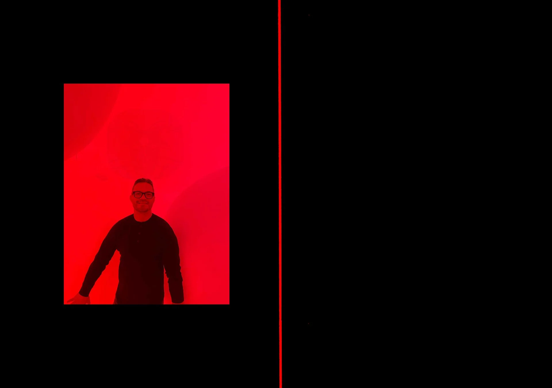 Person standing in front of a red illuminated background, with a black shirt, glasses, smiling and holding a device in one hand.