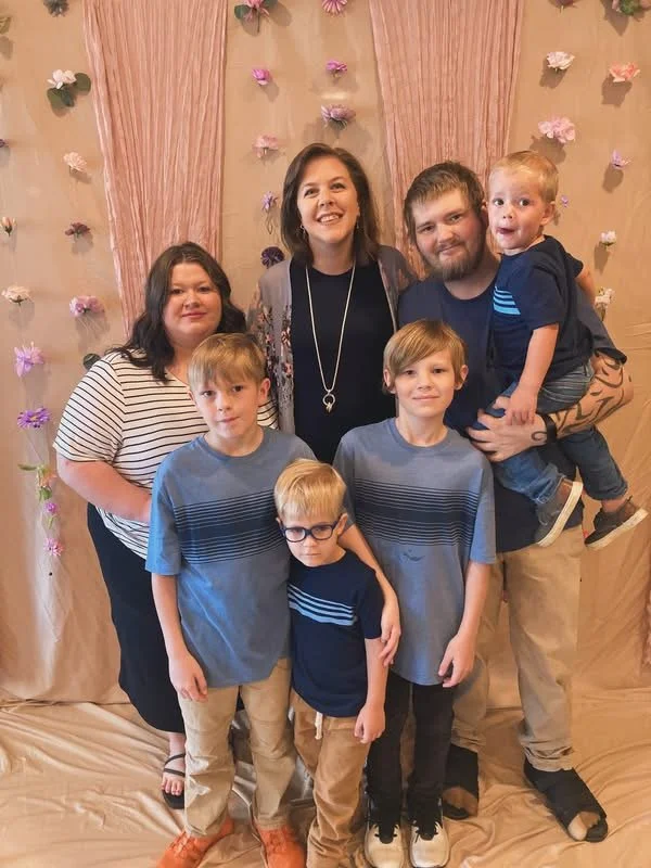 A group of seven people, including children and adults, standing in front of a decorated backdrop with pink and purple flowers and pink drapery, smiling and posing for a photo.