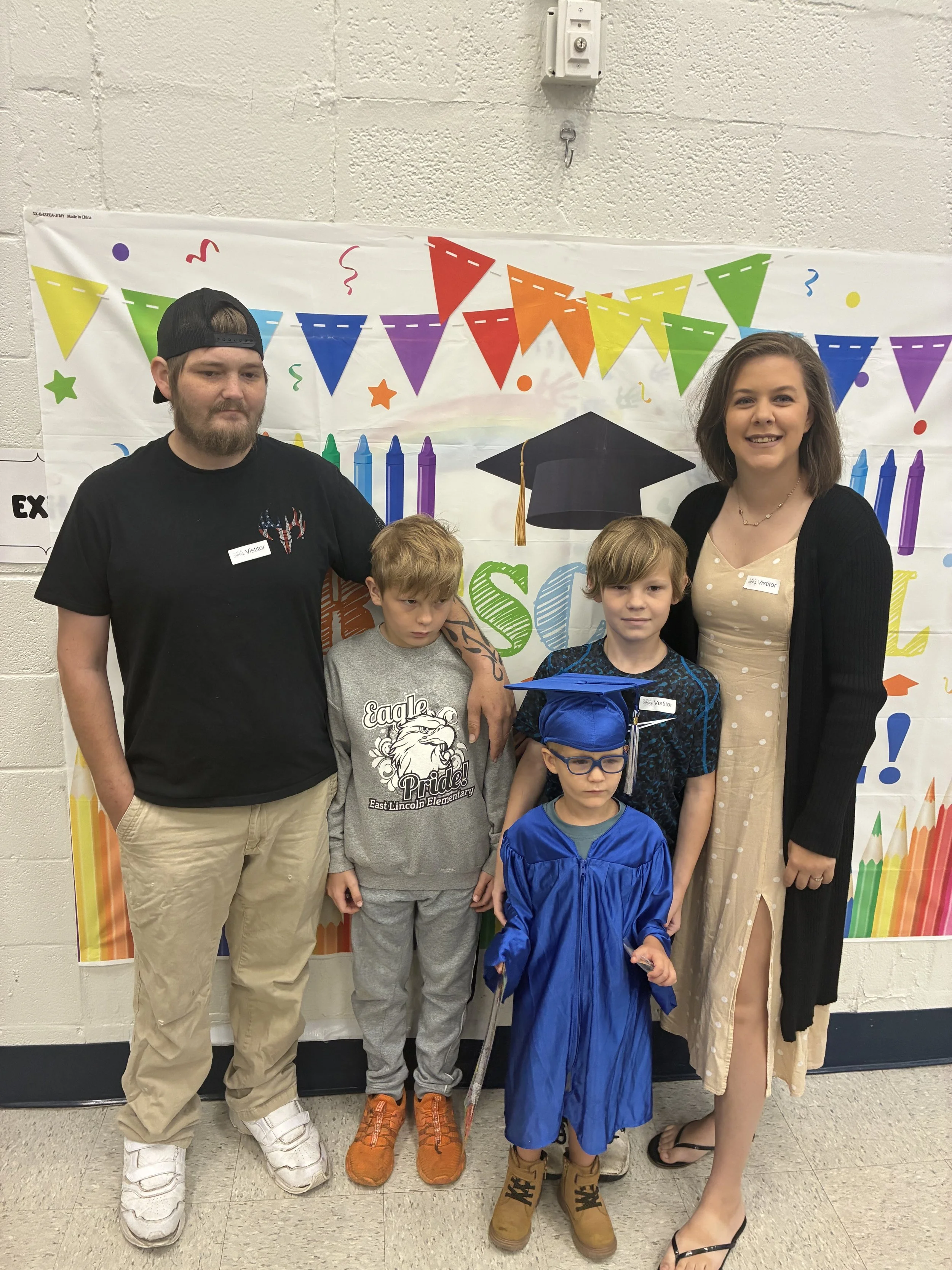 A group of people, including three children and two adults, standing in front of a colorful backdrop with celebrations written on it. The children are dressed in casual clothes and one is wearing a blue graduation cap and gown. The adults are smiling, and the background has a festive design with flags and candles, indicating a celebration, possibly a graduation or school event.