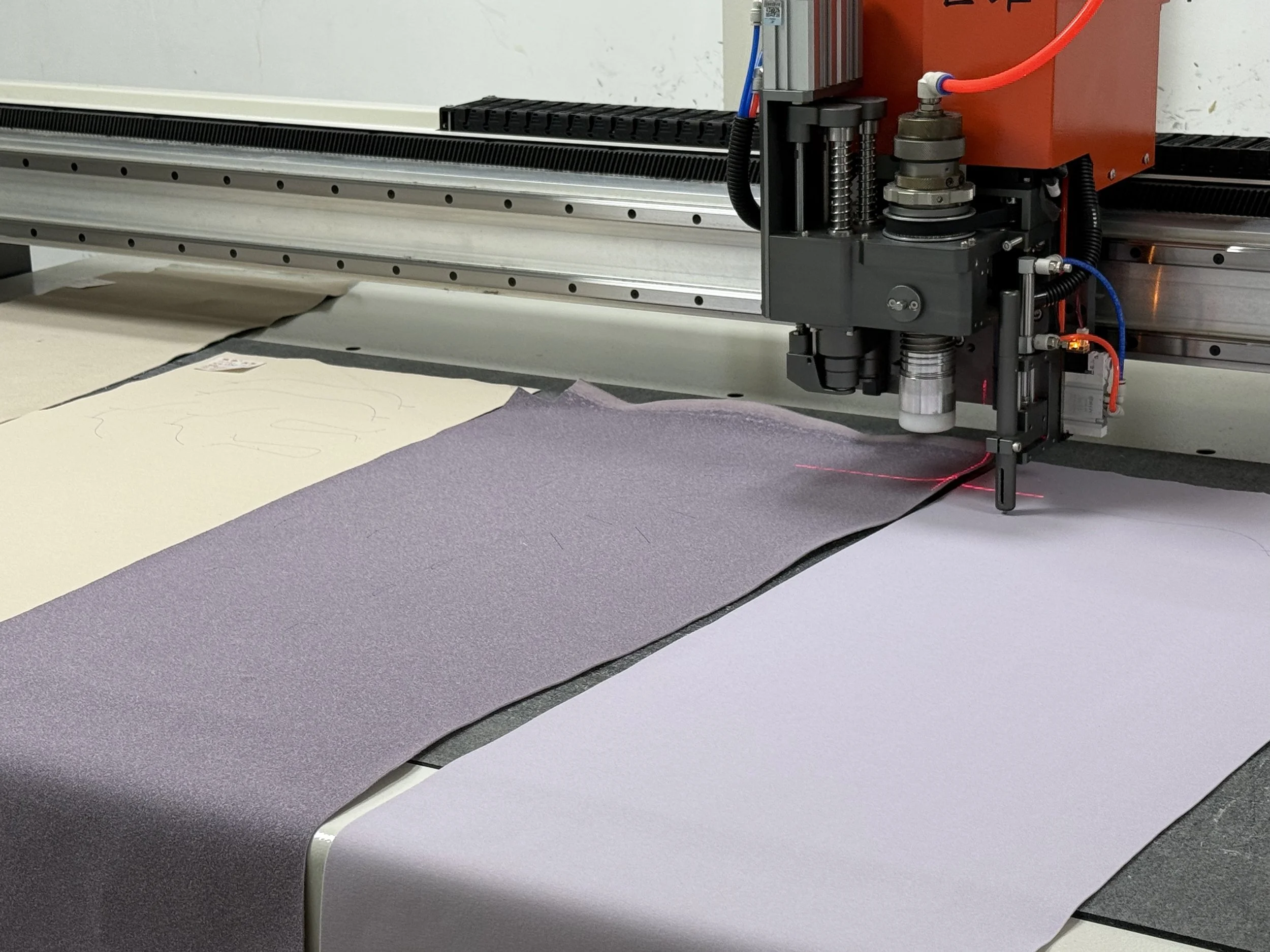 Close-up of a CNC machine working on a sheet of colored fabric or material. The machine's tool emits a red laser beam, and various mechanical parts and wires are visible on the right side of the image.