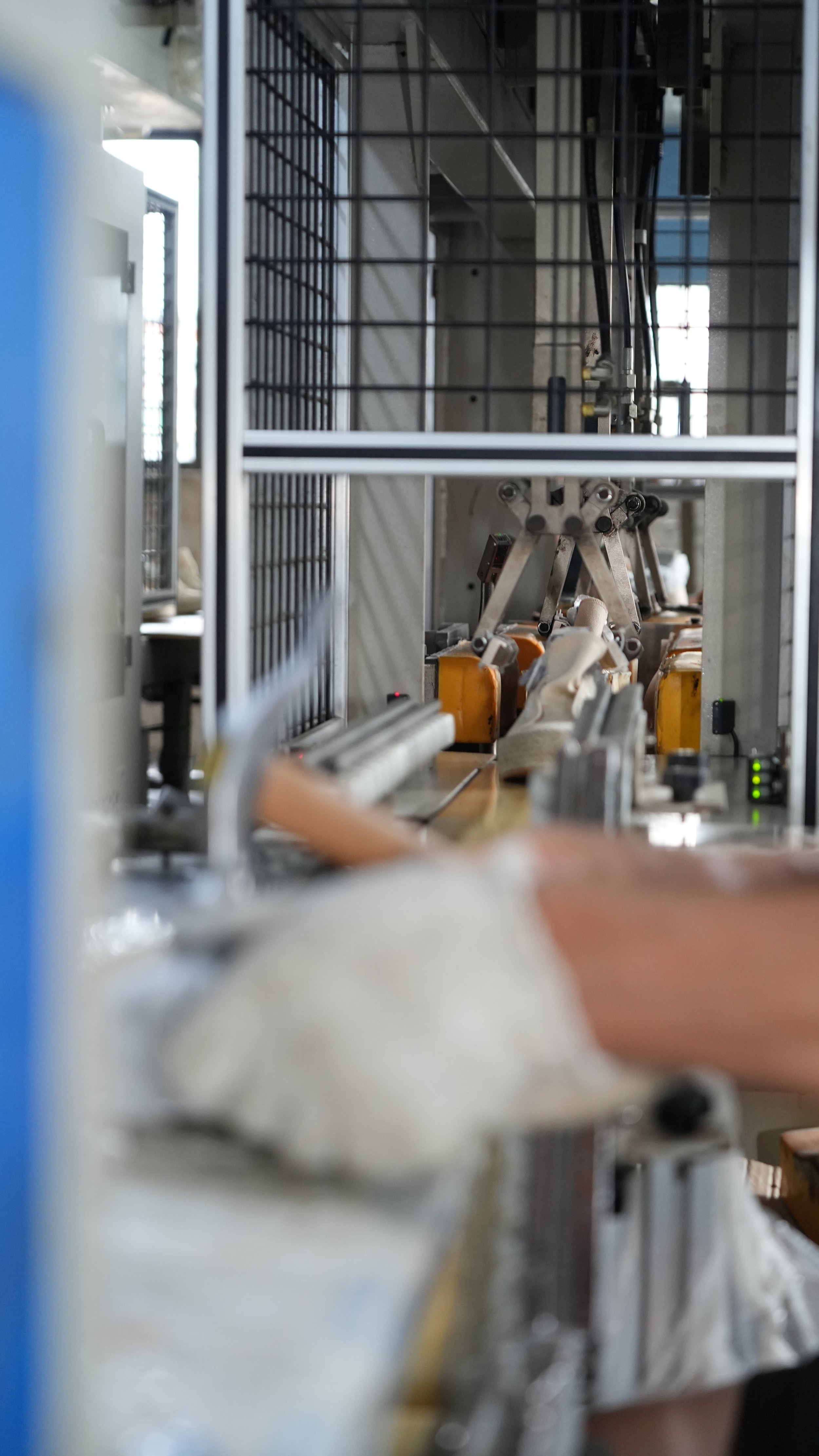 Close-up view of a robotic arm in an industrial setting, working on a conveyor belt with shoes, surrounded by metal framework and machinery.