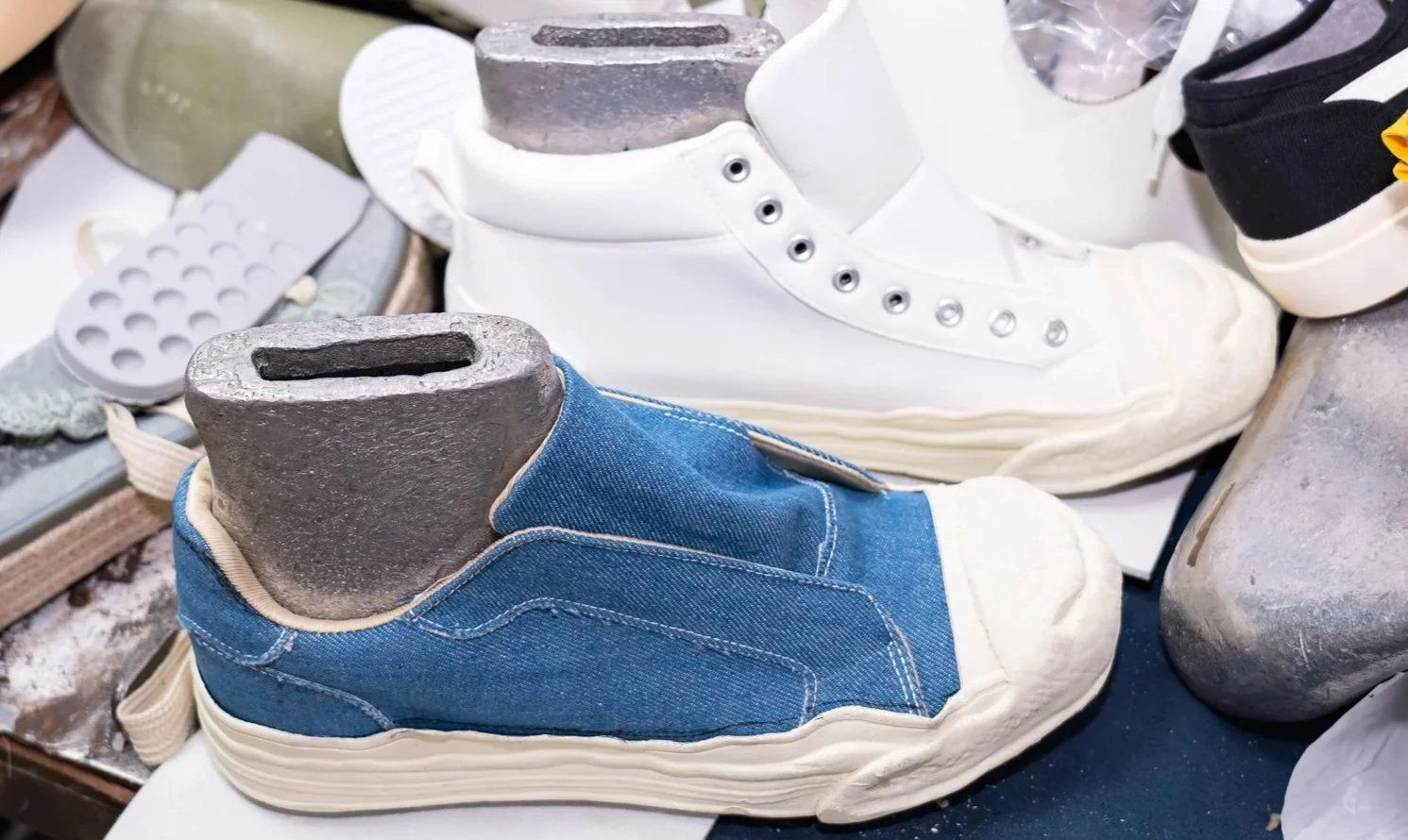 Pair of shoe molds, one in denim fabric and the other in white, surrounded by various shoe components and tools.