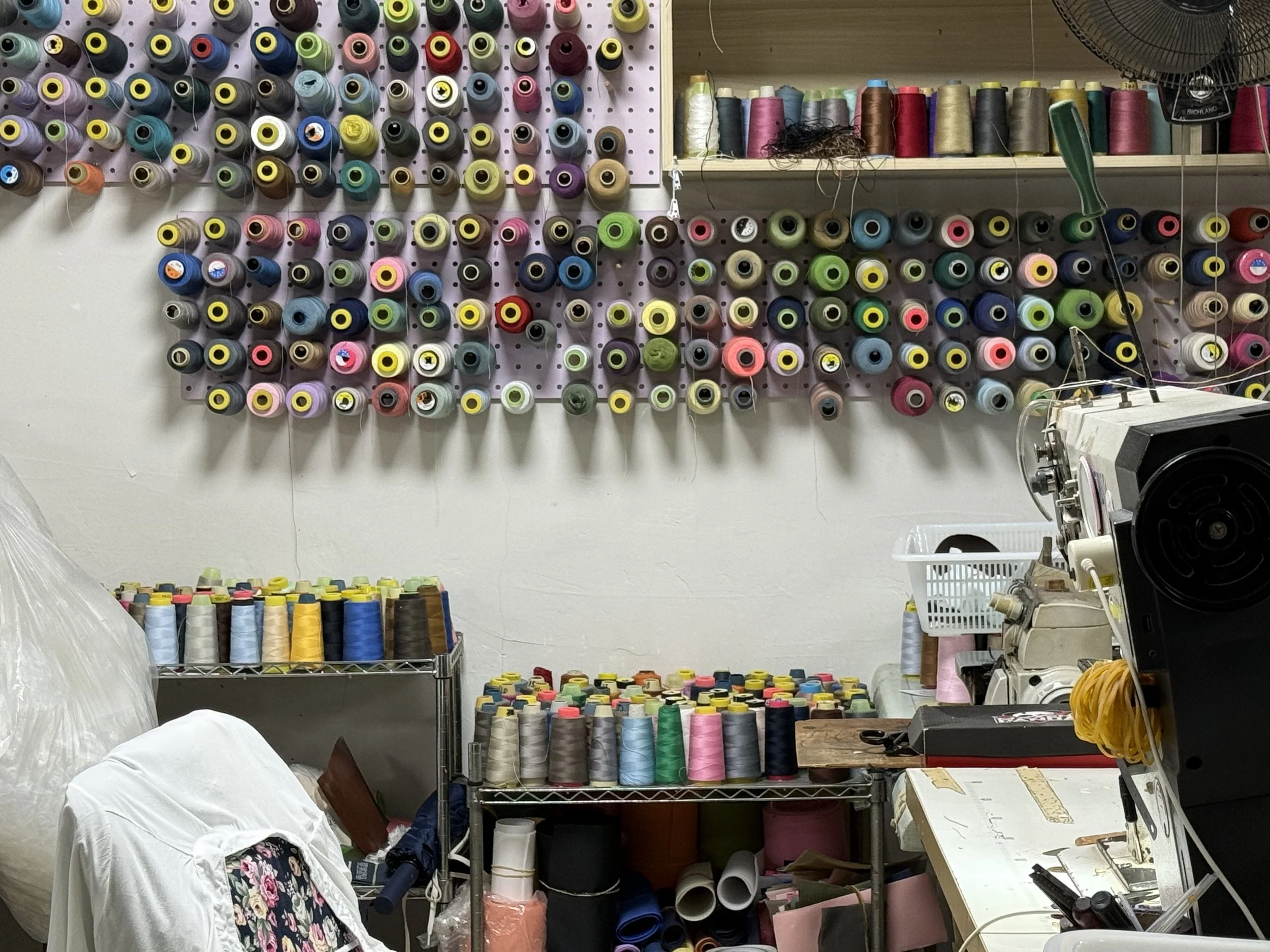 Wall with numerous spools of colorful thread arranged in rows, some stored on shelves and others hanging on hooks, within a sewing or textile workshop.