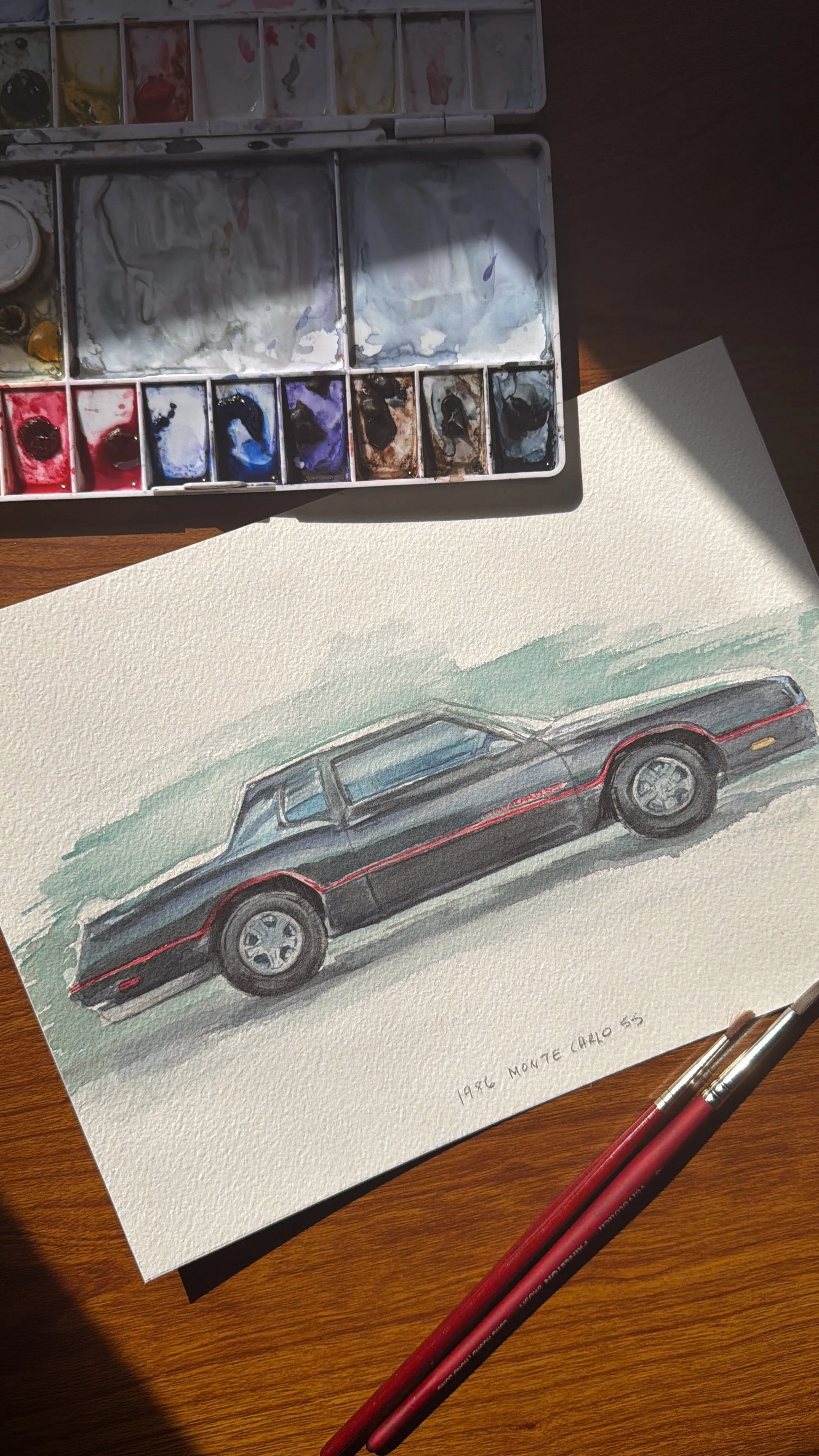 A watercolor painting of a 1956 Monte Carlo SS car on white watercolor paper, with watercolor paints and brushes on a wooden surface nearby.