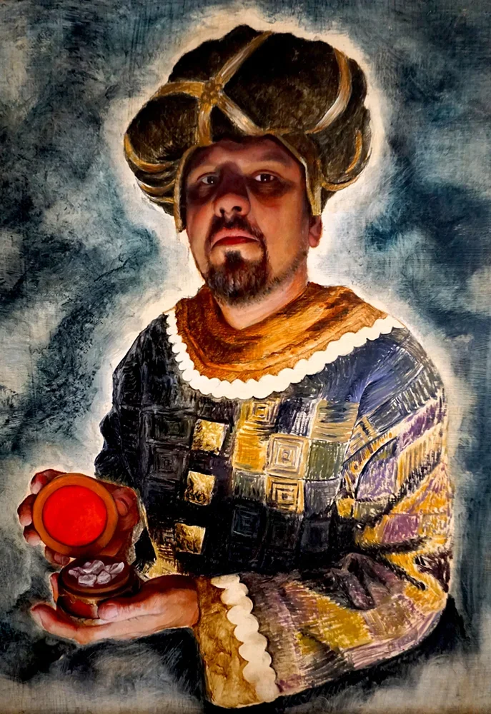 A man with a beard and mustache dressed in a patterned robe, wearing a traditional headdress, holding a red box with a lid, in front of a painted background.