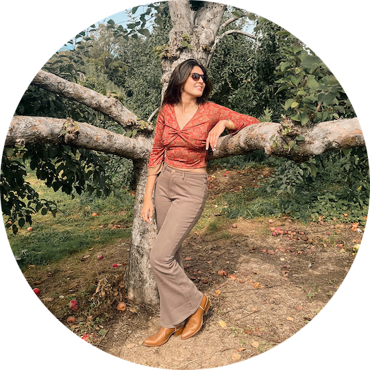 A woman wearing sunglasses, a red patterned blouse, beige wide-leg pants, and tan boots standing outdoors next to a tree with sprawling branches.