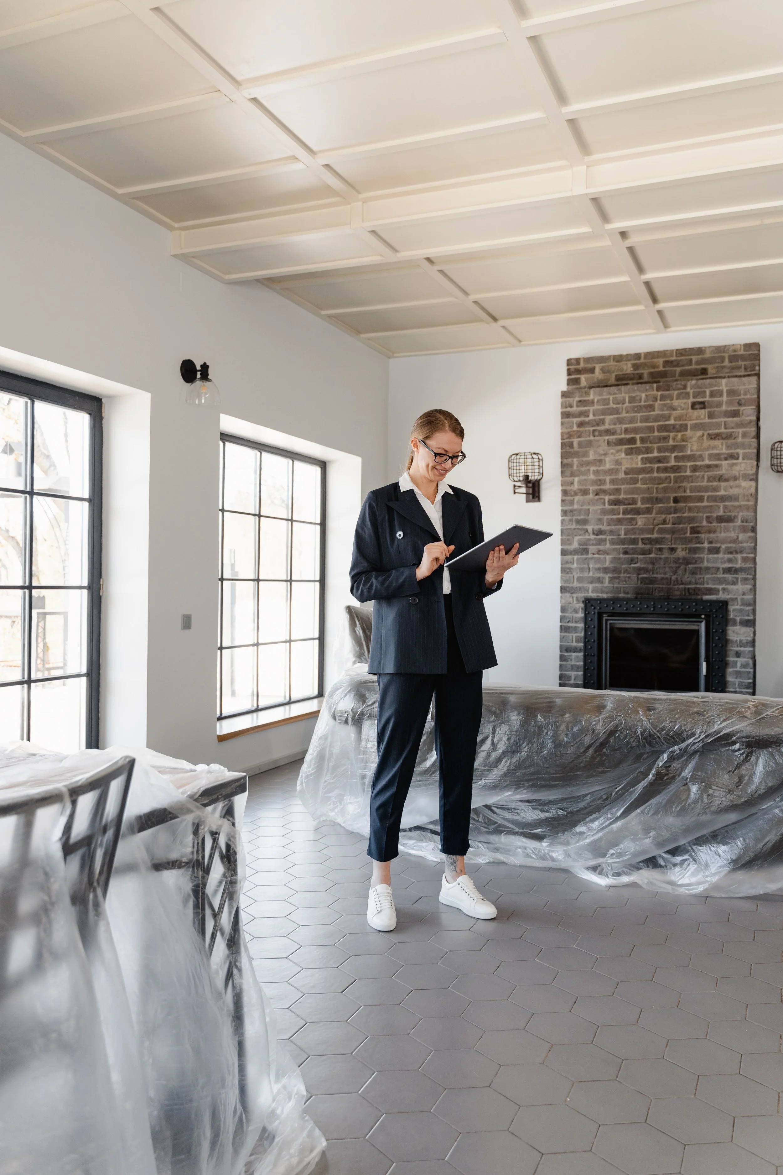 A woman with glasses in business attire, wearing white sneakers, standing in a modern living room with large windows, a brick fireplace, and a vaulted ceiling, looking at a tablet device.