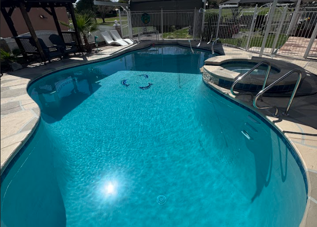 An outdoor swimming pool with clear blue water, surrounded by a concrete deck, white lounge chairs, and a safety fence. There is a hot tub or spa attached to the pool, accessed by metal handrails.