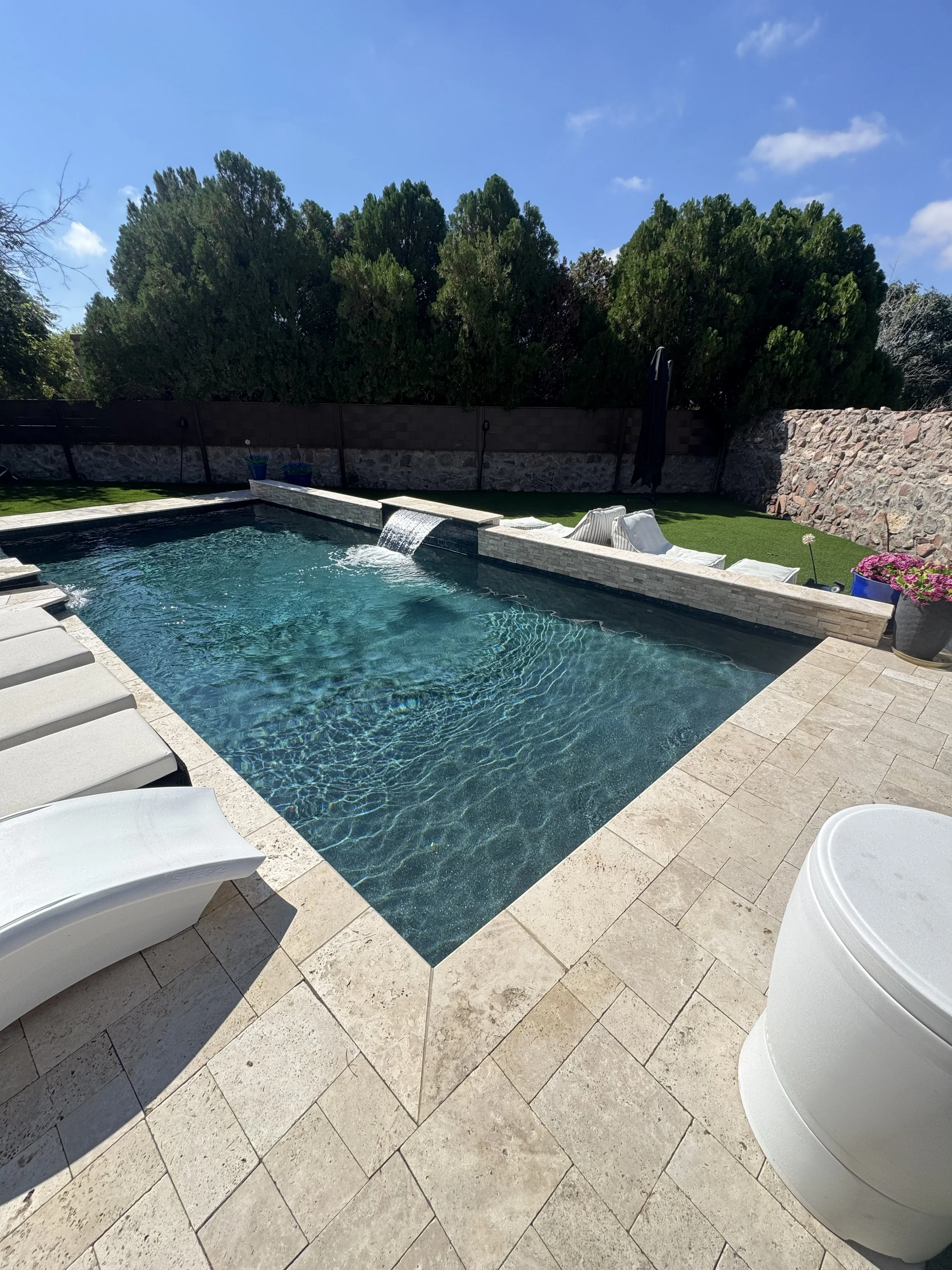 A backyard with a swimming pool, poolside chairs, a black umbrella, potted plants, and a stone wall with dense trees against a blue sky.