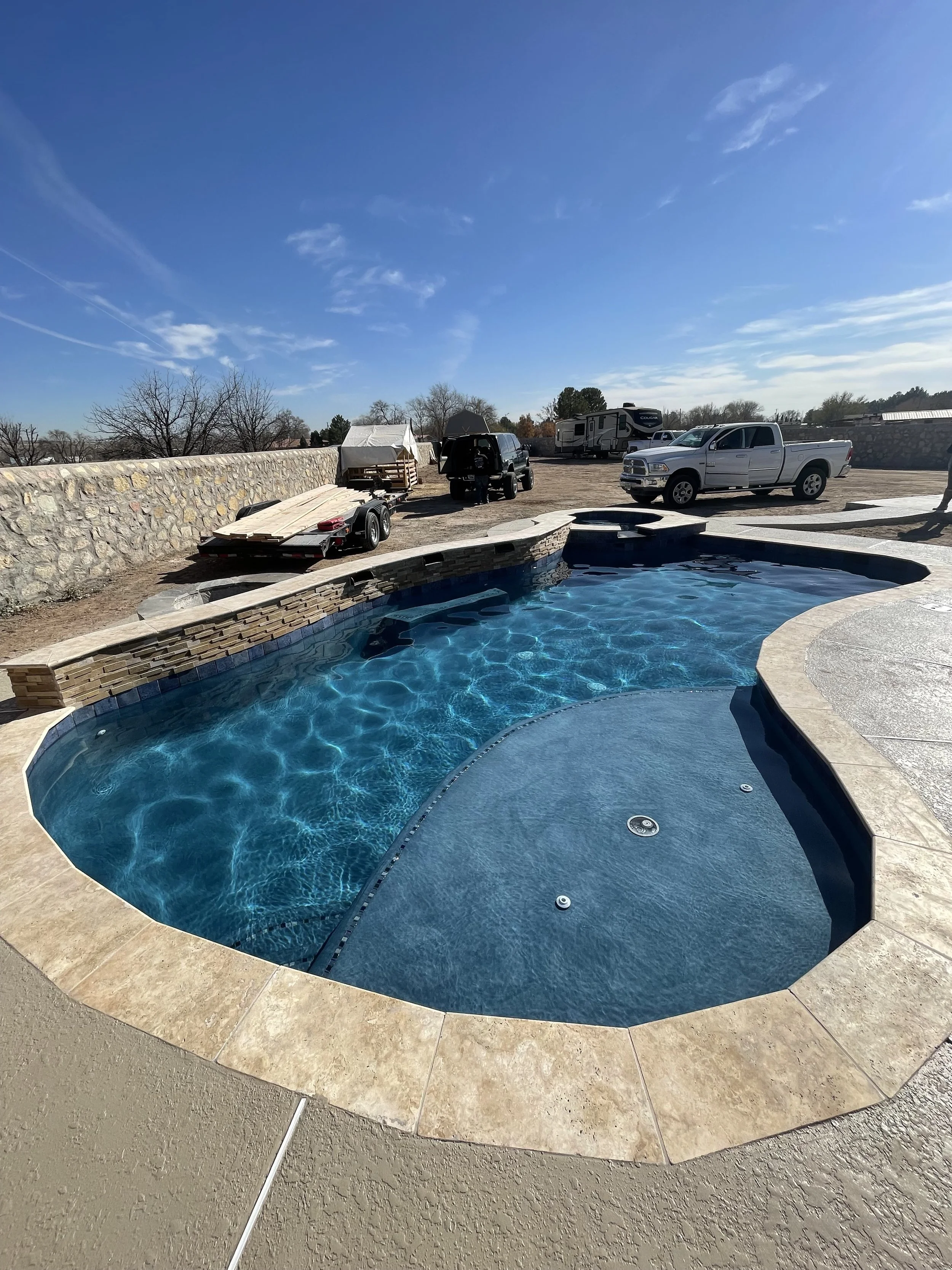A backyard with a swimming pool, a stone wall, a trailer with wood planks, several vehicles including trucks, and RVs. The sky is mostly clear with a few clouds.