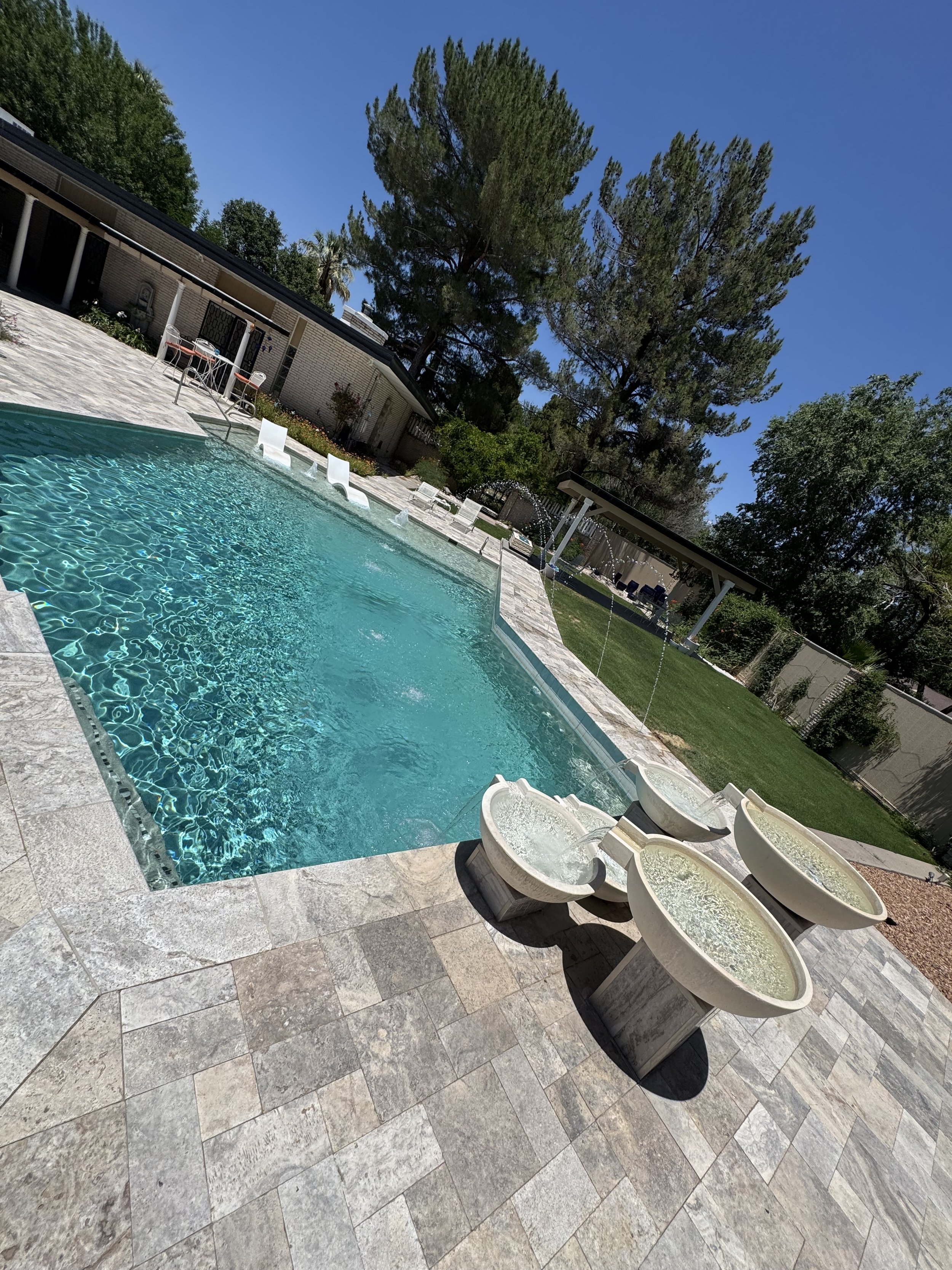 Backyard swimming pool with a tiled deck, surrounded by green trees and bushes under a clear blue sky, with poolside chairs and fountains.