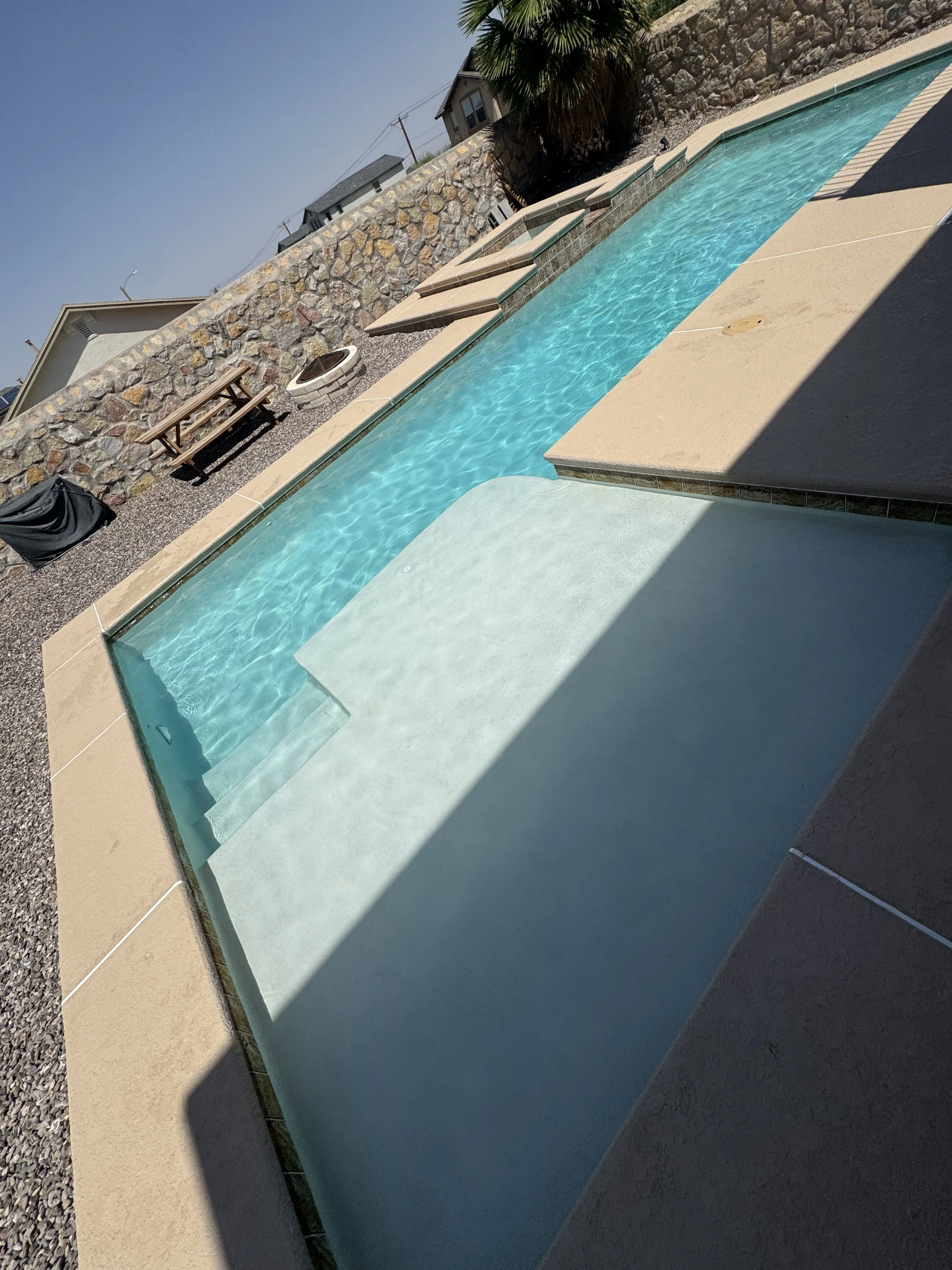 A backyard swimming pool with turquoise water, beige pool deck, and a stone wall in the background. There is a wooden picnic table and a fire pit on the gravel area, with a tall palm tree nearby. Houses and power lines are visible beyond the wall.