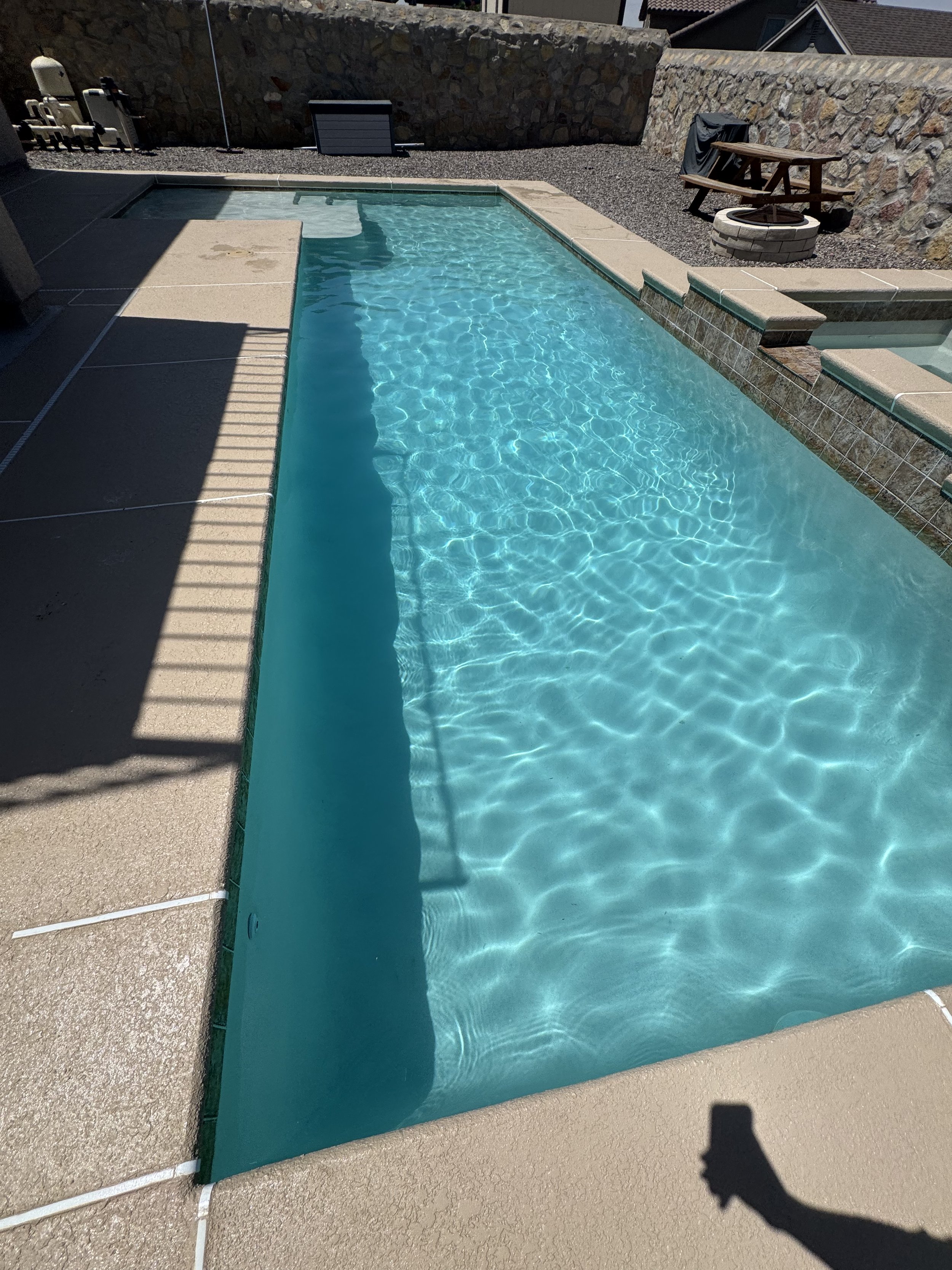 A backyard with a swimming pool, surrounded by concrete and stone walls, with a small fire pit and a picnic table in the background.