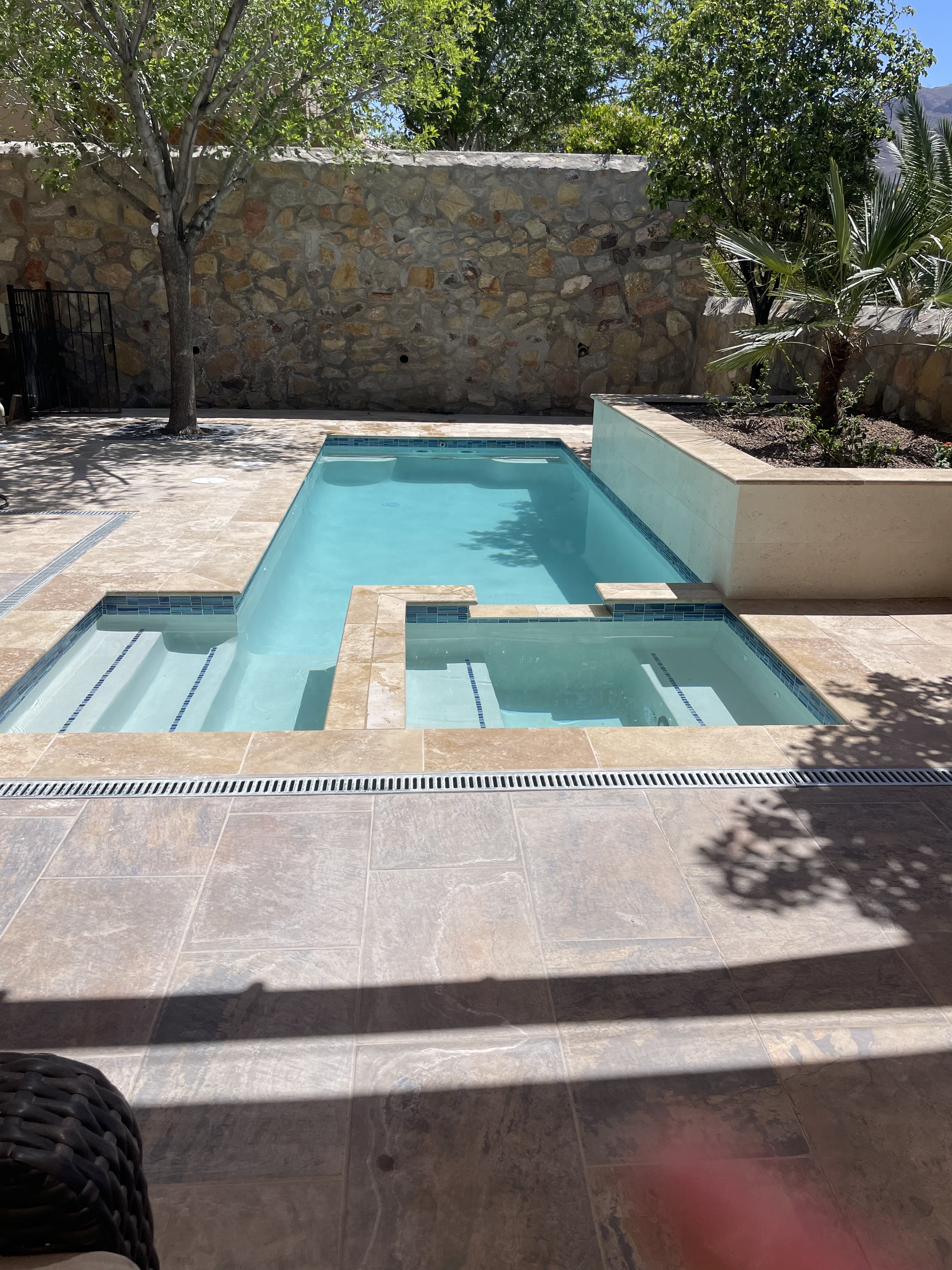 A small outdoor swimming pool with beige tiles, a black metal gate, and a stone wall in the background. There are trees and plants around the pool area, with some shadows cast on the floor.