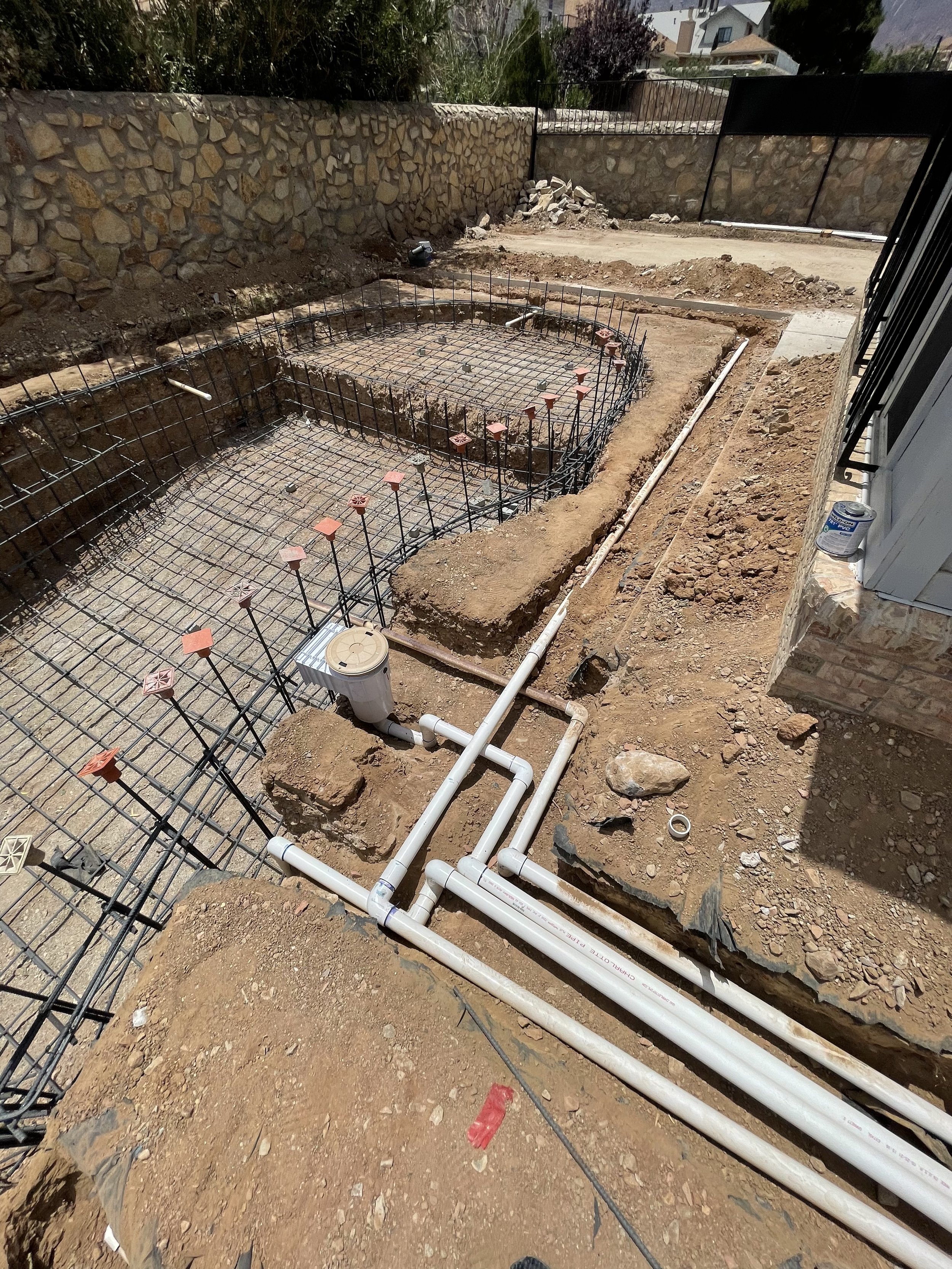Construction site with foundation reinforcement and plumbing pipes in ground, stone wall in background.