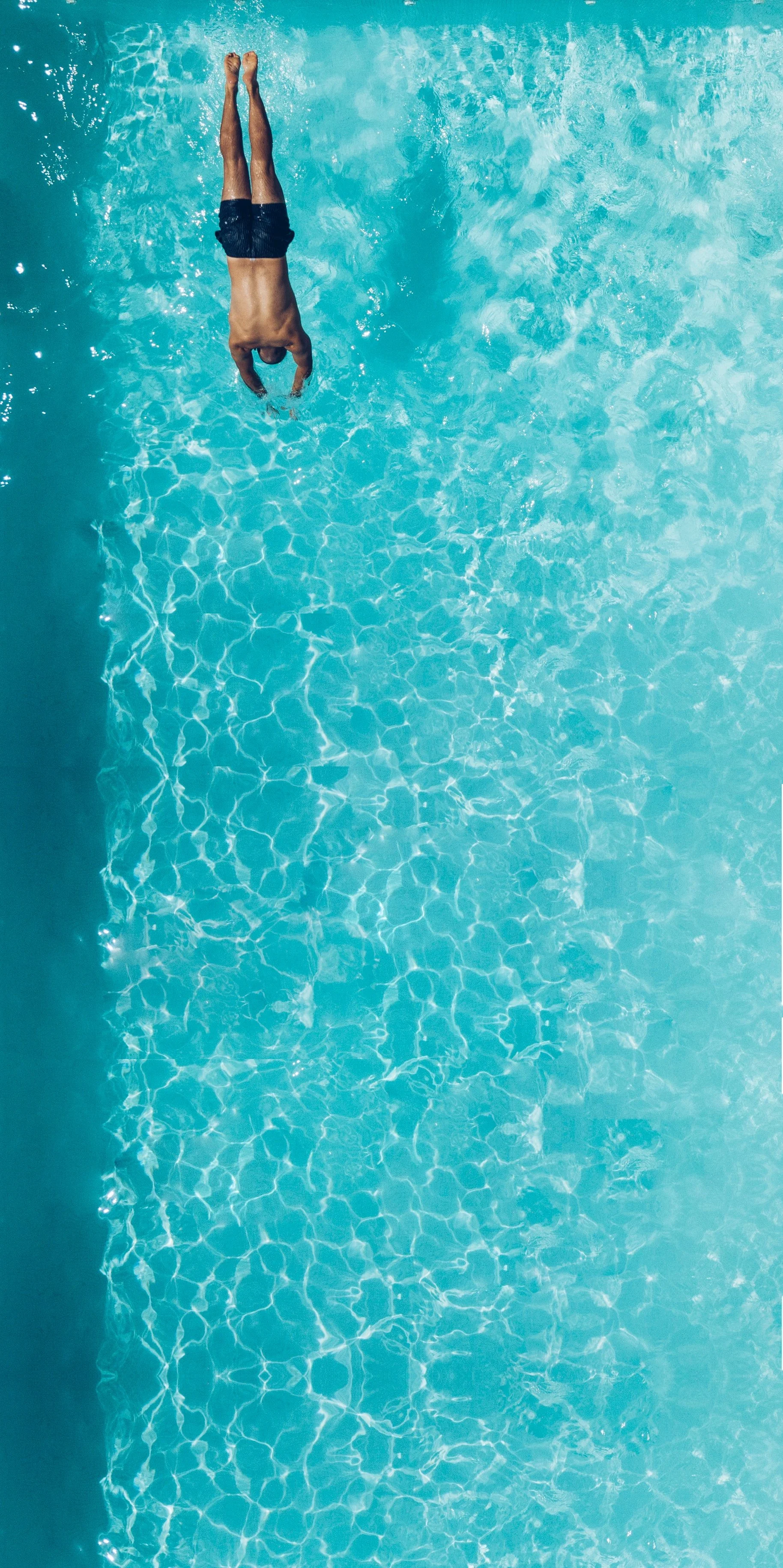 A person diving into a light blue swimming pool, wearing black swimming trunks, viewed from above.