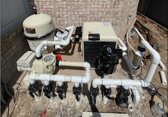 An outdoor swimming pool pump system with various pipes, filters, and valves, installed on a concrete surface next to a brick wall.