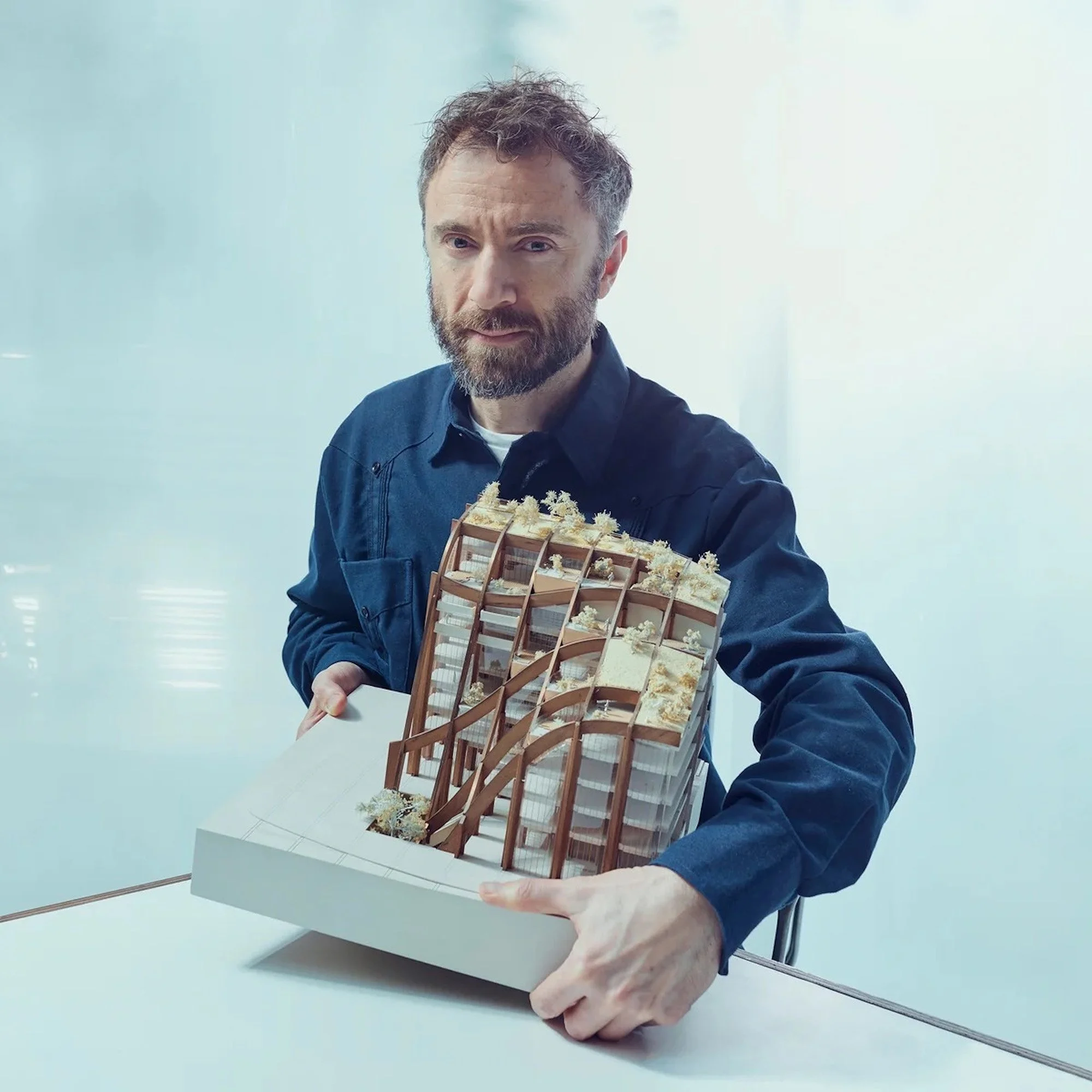 A man with a beard and mustache holding a model of a multi-story building during a presentation or display.