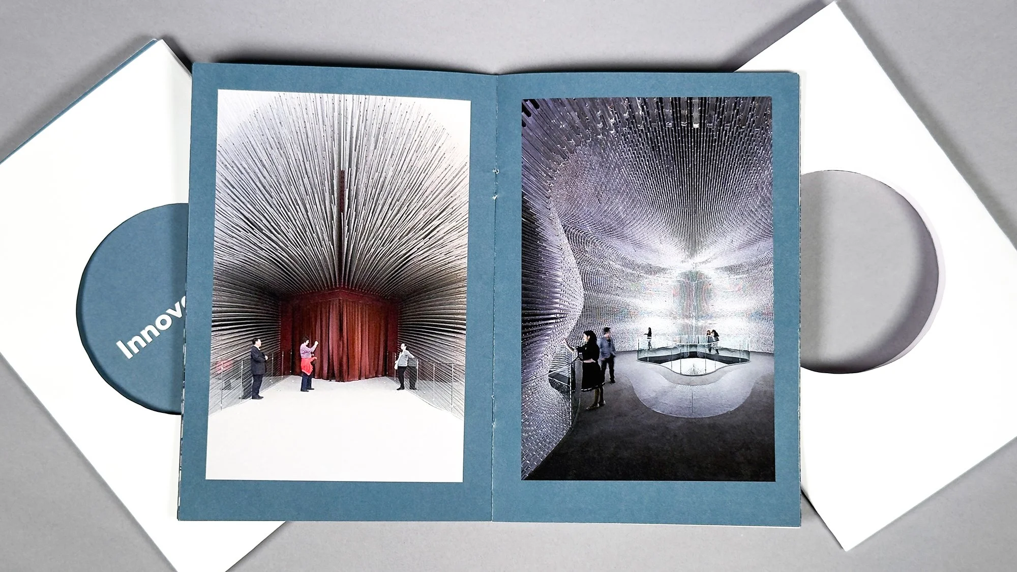 Open booklet showing interior designs of the Seed Cathedral, appearing as modern architectural structures with visitors inside, placed on a white surface.