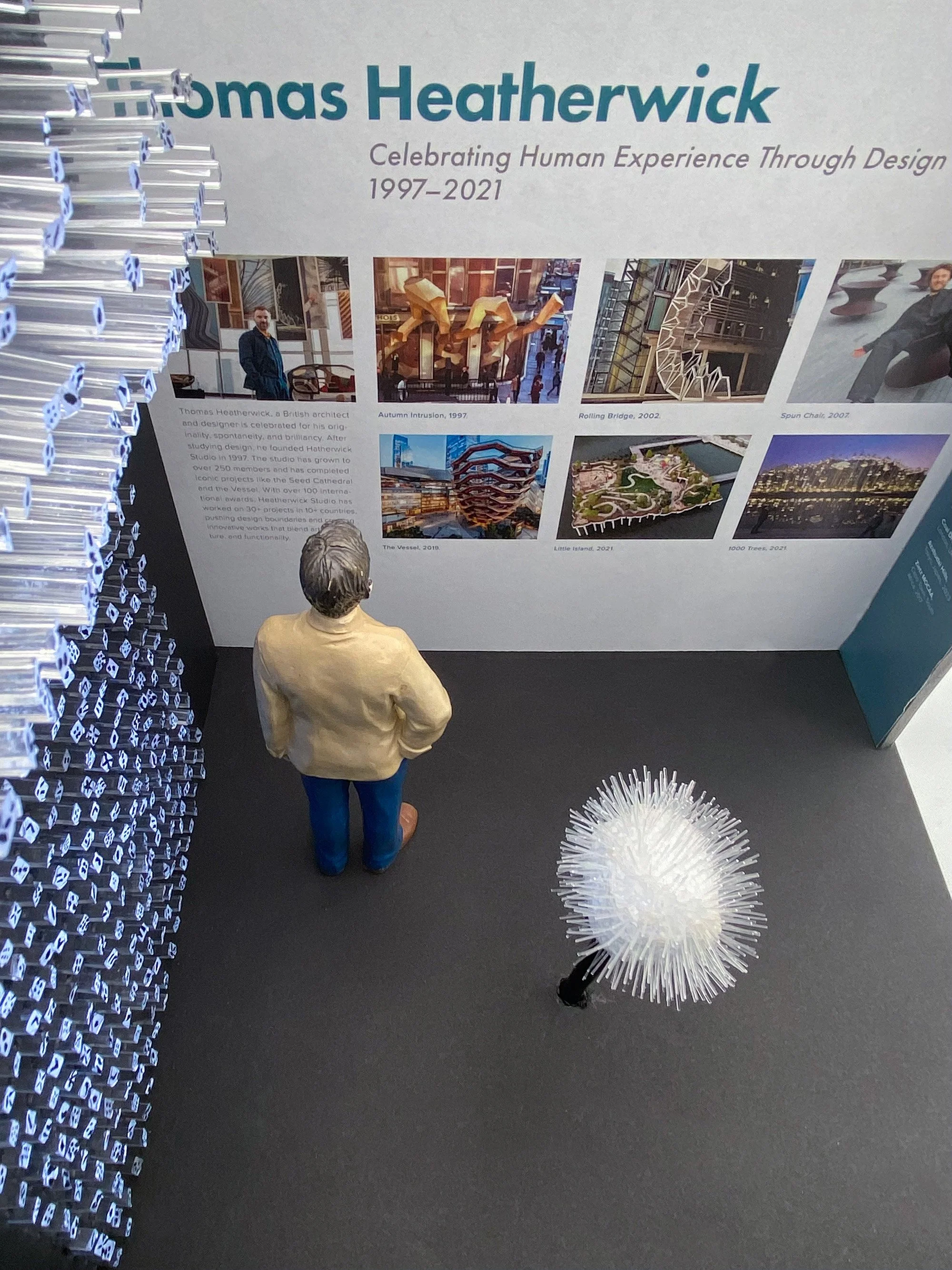 Display board with images of architectural projects and a small figurine of a man in a beige jacket looking at it while staring next to acrylic rods protruding the left wall, and to his right is a small spiky dandelion-like sculpture.