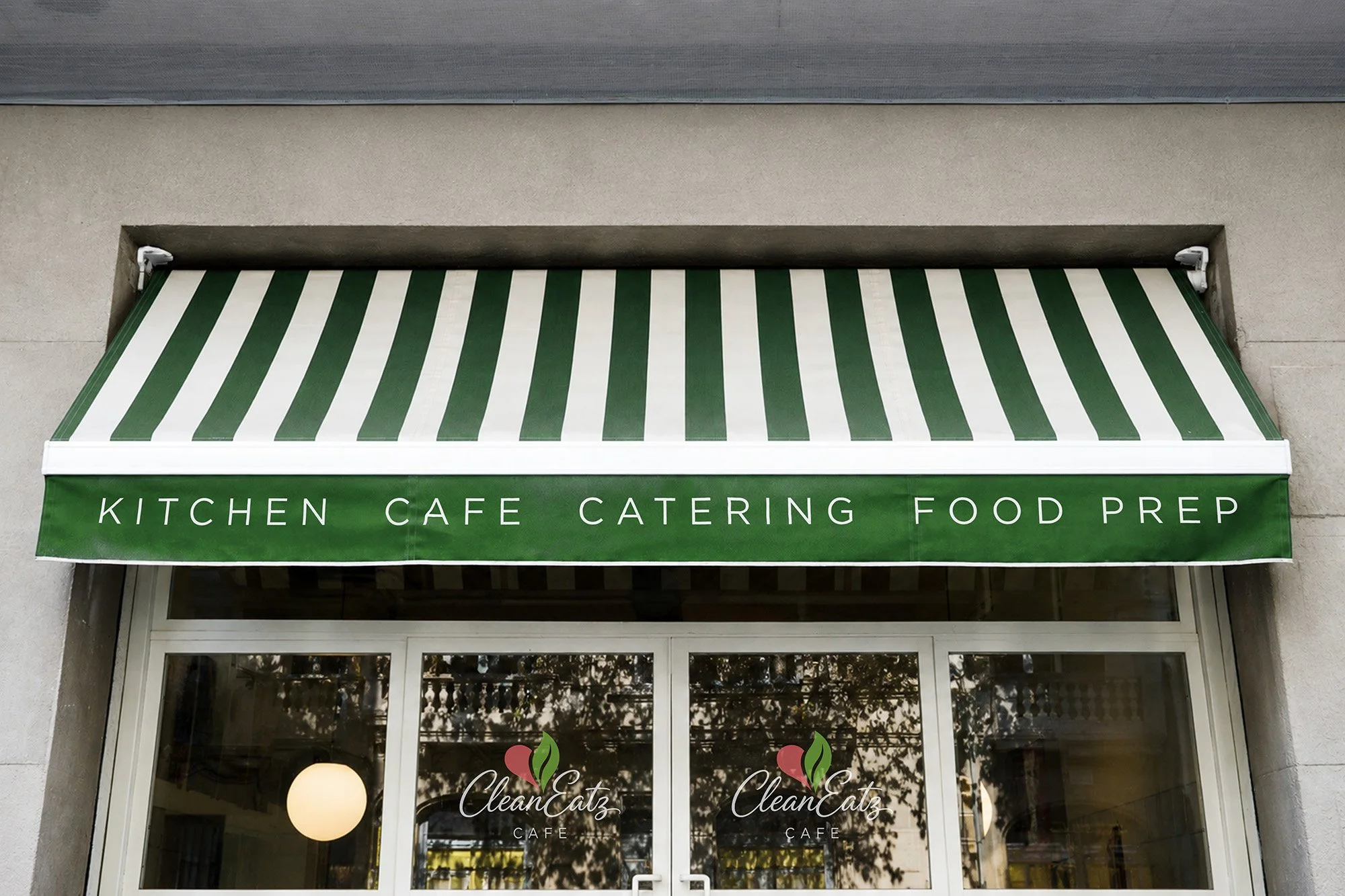 Green and white striped awning over a cafe entrance with the words 'KITCHEN CAFE CATERING FOOD PREP' written on it. The entrance has glass doors with the café's logo, 'CleanEatz Cafe.'