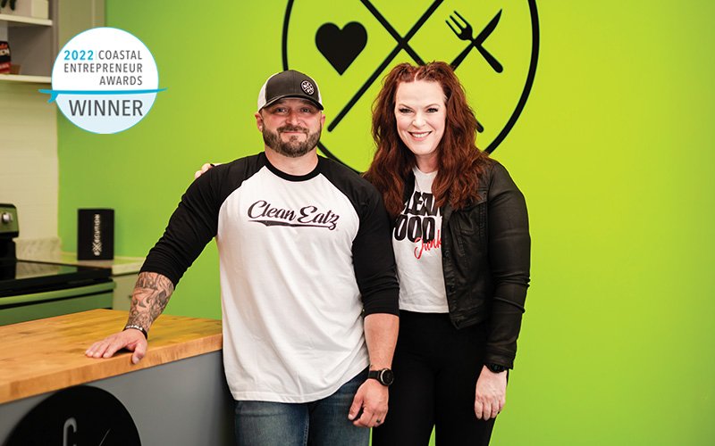 A man and a woman standing together indoors, smiling at the camera. The man is wearing a baseball cap, a white and black baseball tee with 'Clean Eatz' on it, and has tattoos on his left arm. The woman has red hair and is dressed in a black jacket over a graphic t-shirt. They are standing in front of a bright green wall with a logo and a badge on it that indicates they are winners of the 2022 Coastal Entrepreneur Awards.