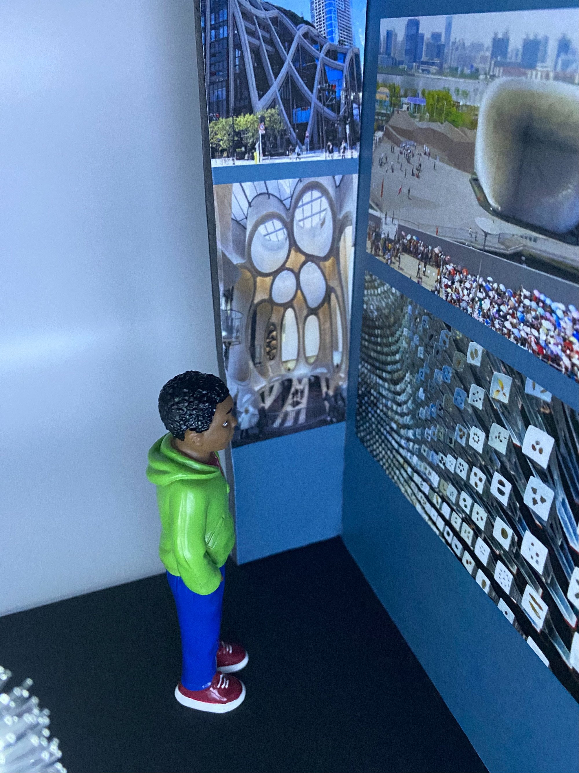 A child-sized mannequin wearing a green hoodie and blue pants, with red and white sneakers, is standing in front of a display of close up images of the interior of the seed cathedral other images include glass artwork with circular window designs.