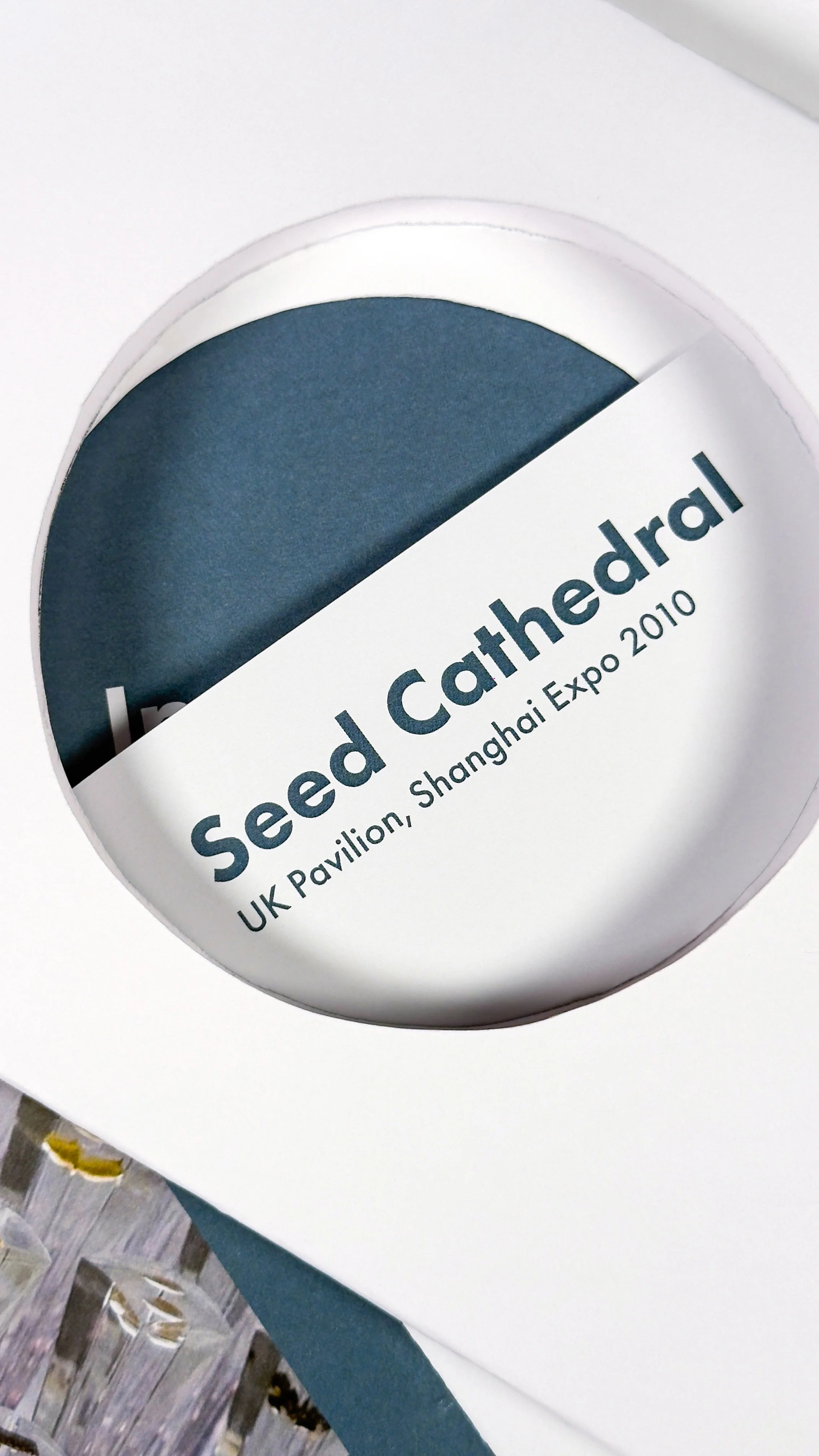 Close-up of a circular cutout looking through a section of the book called 'Seed Cathedrals UK Pavilion, Shanghai Expo 2010' on a white background and partially visible dark blue section.