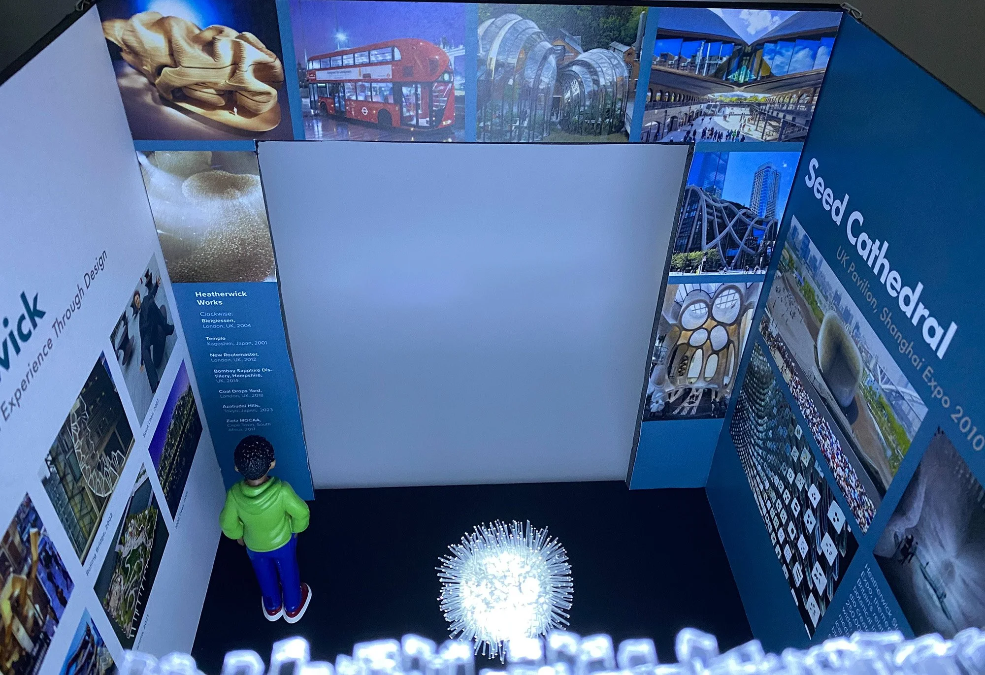 Display board showcasing various architectural designs and projects, including a wooden sculpture, a red double-decker bus, glass greenhouses, futuristic buildings, and a cityscape. Two other walls expand on the artist and the Seed Cathedral.