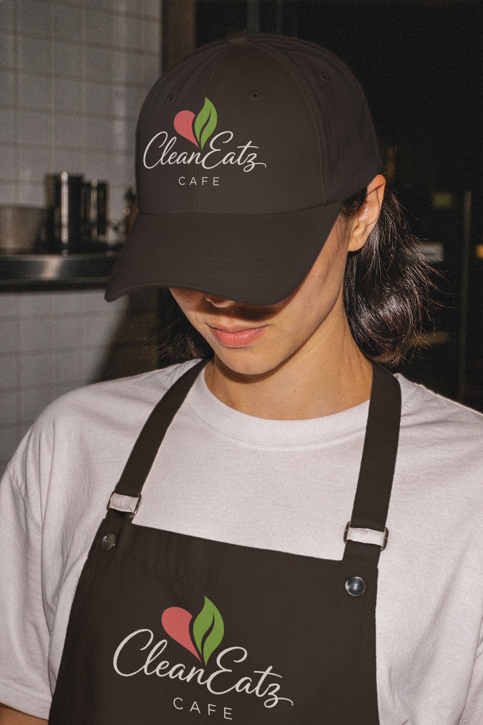 A person wearing a black cap and apron with 'Clean EatZ Cafe' logo, standing in a kitchen.