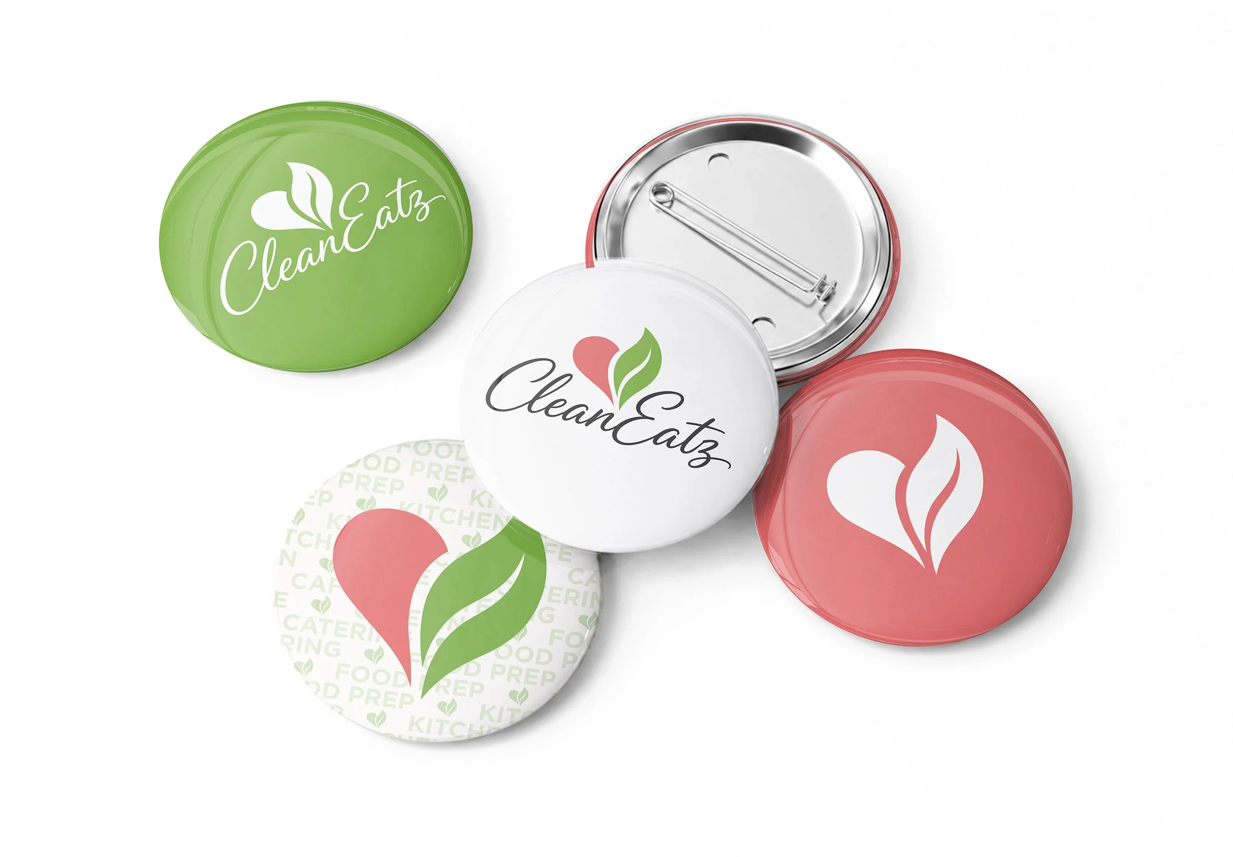 The image shows several pins, featuring a green, red, and white color scheme with the logo 'CleanEatz' and a heart-leaf design. Some pins are upside-down, revealing metal interior lids with a latch.
