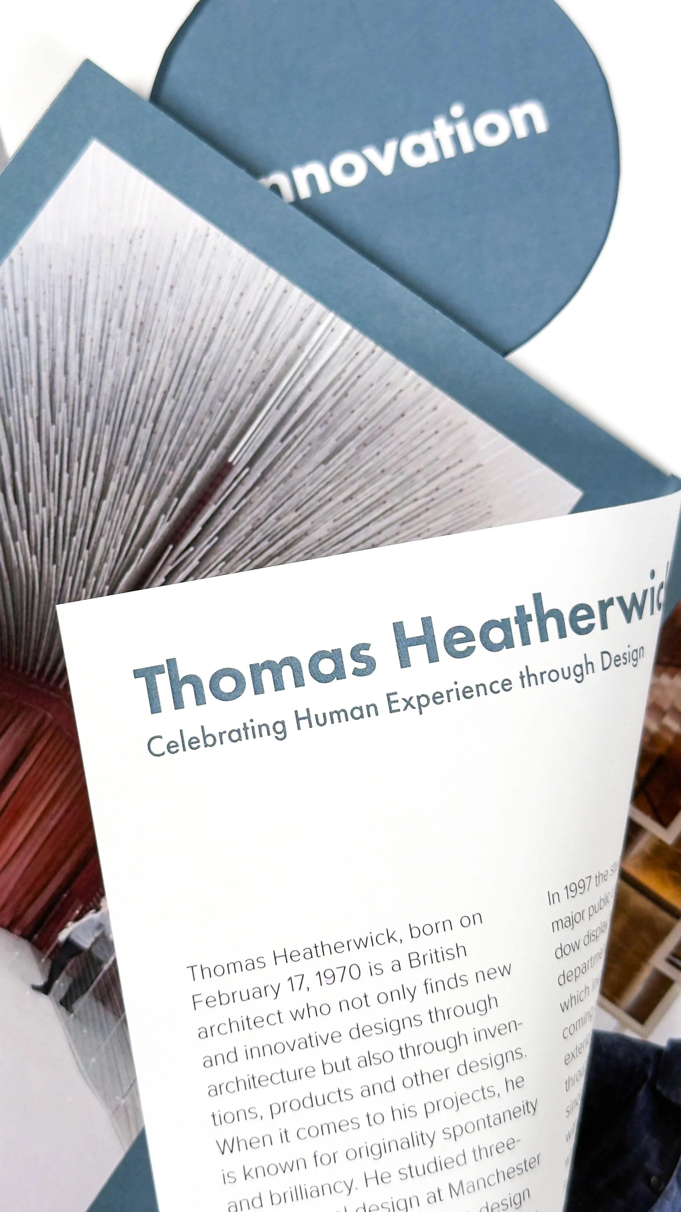 A close-up photograph of a brochure with a page flipping that reads 'Thomas Heatherwick', featuring the words 'Celebrating Human Experience through Design'. Behind the page is an image of the entrance to the Seed Cathedral.