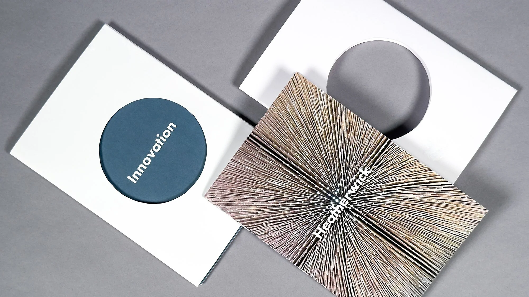 Three sheets of paper with different designs, one with a blue circle cut out and the word 'Innovation', one with a radial burst struct with the word 'Heatherwick', and one with a geometric cutout and circular hole, all placed on a gray surface.