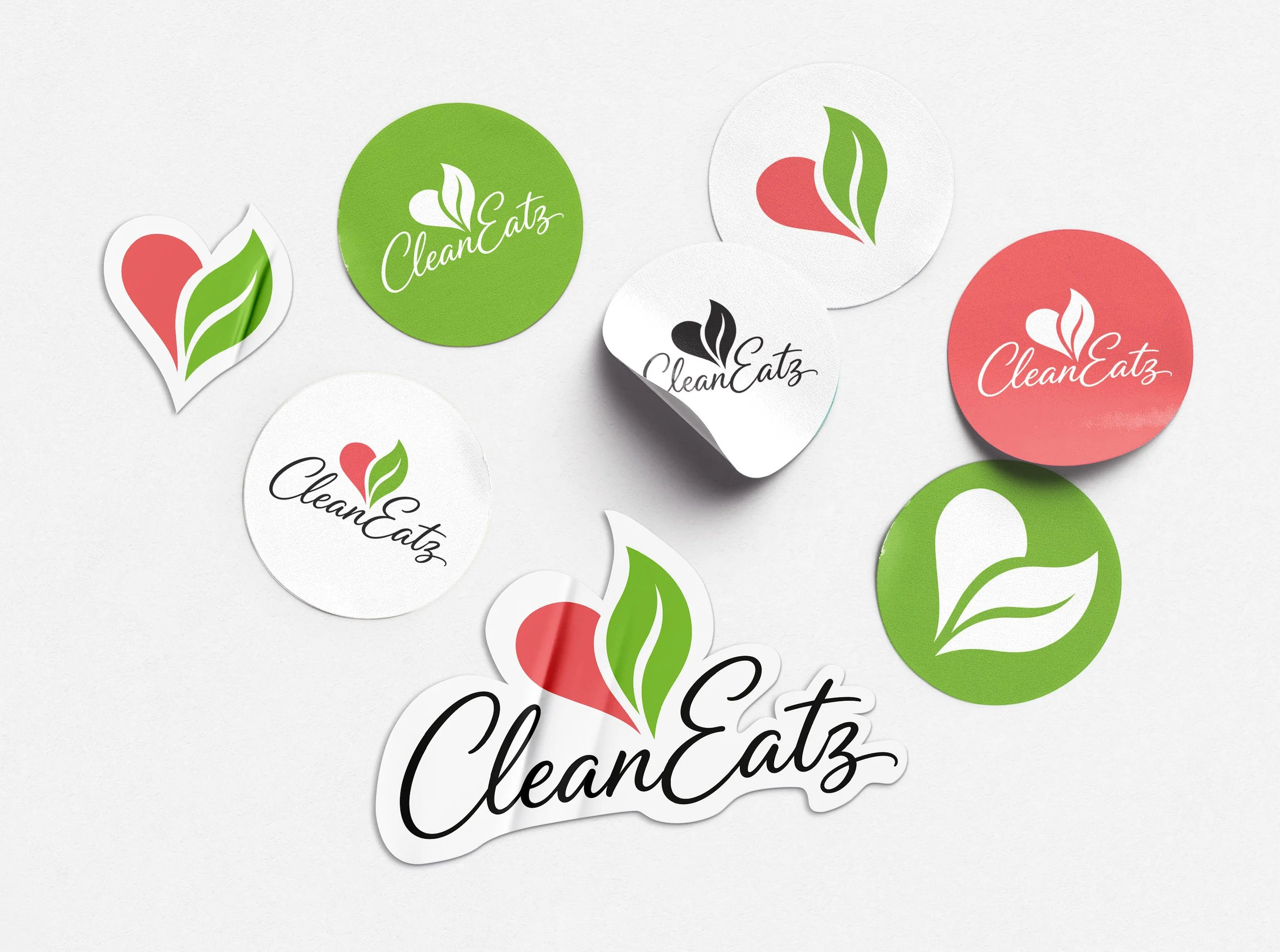 Circular and stylized shaped stickers with the logo 'CleanEatz' that features a heart-shaped leaf and stylized text, in green, white, black, and red, scattered on a white surface.