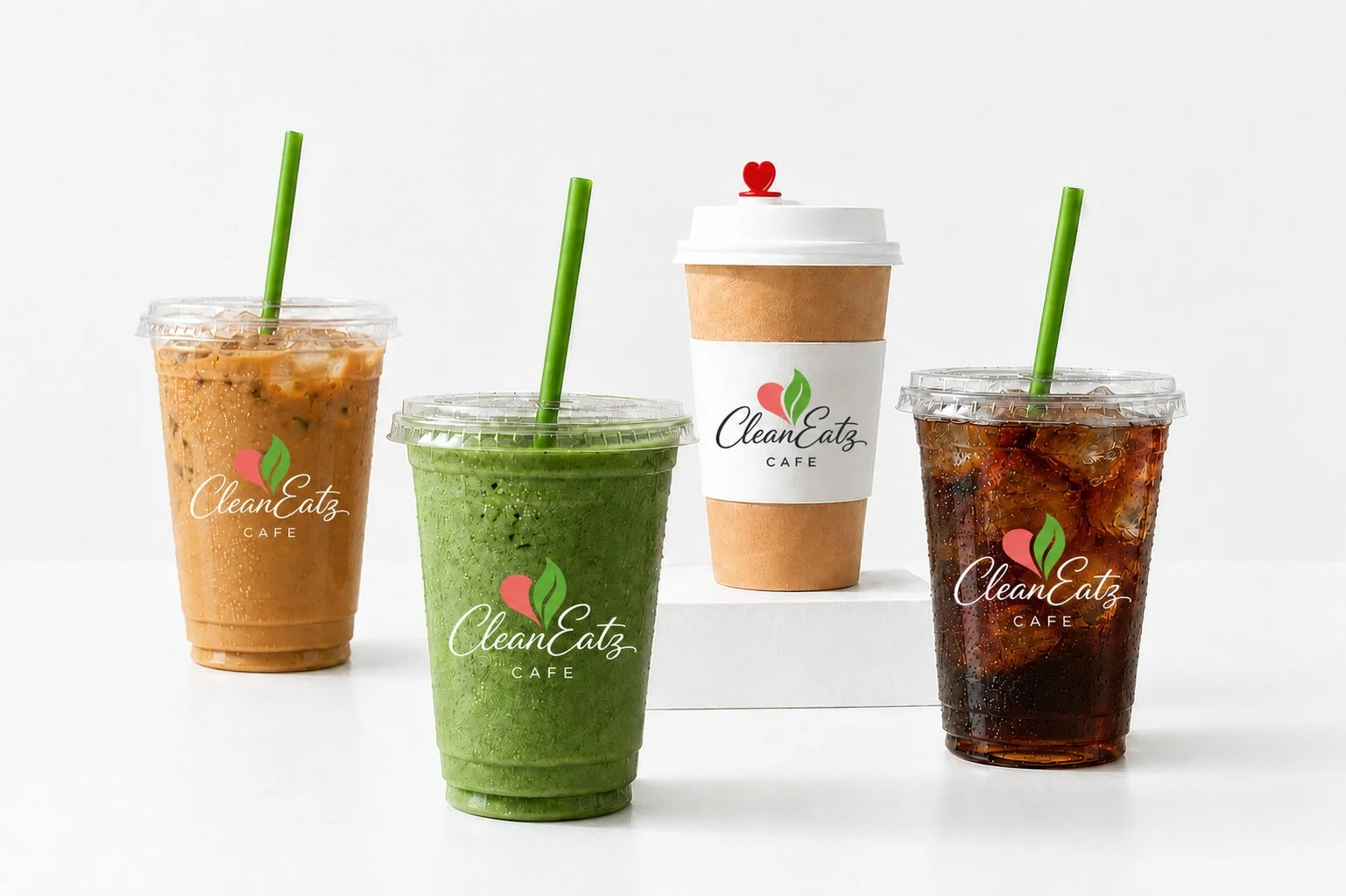 Four drinks from CleanEatz Cafe, including three iced beverages, a coffee, a  matcha and a soda, in plastic cups with green straws, a hot beverage in a paper cup with a sleeve and a lid, all with the cafe's logo, on a white background.