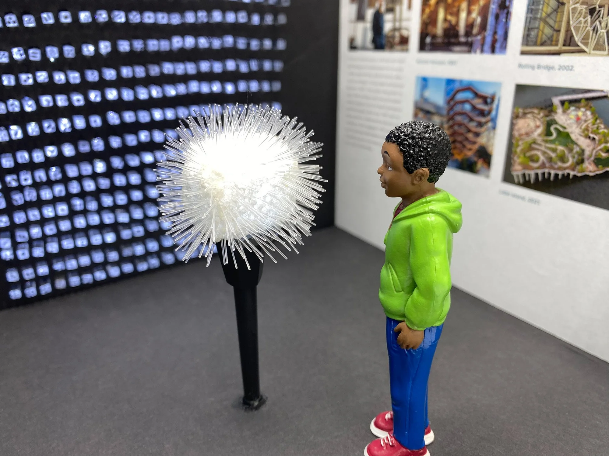 A small figurine of a boy with curly black hair, wearing a green hoodie and blue pants, standing on a black surface. In front of the figurine, there is a bright, spiky light fixture creating a starburst effect. The background features a wall with illuminated icons on the left and a display board with pictures and text on the right.
