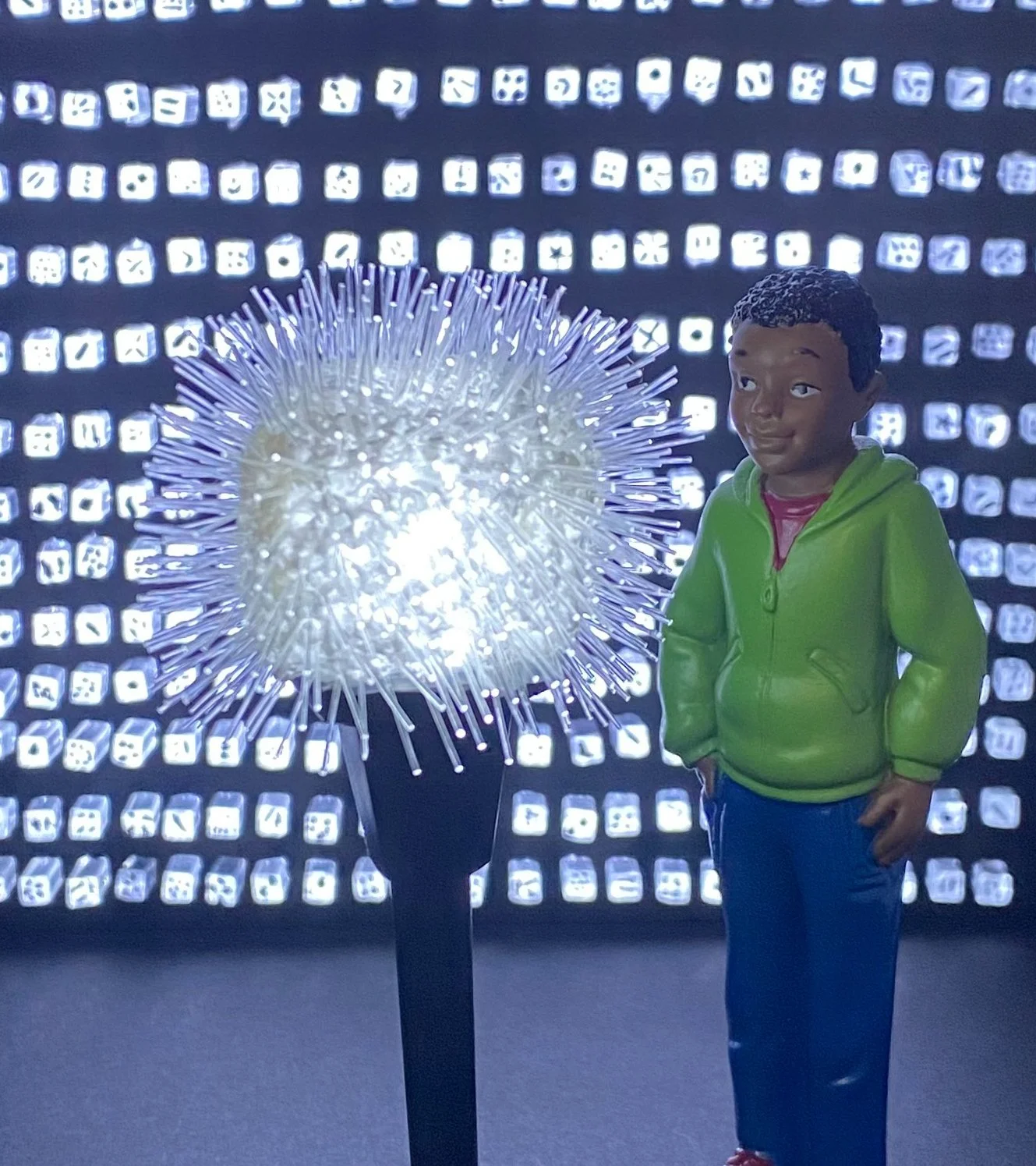 A boy wearing a bright green jacket standing next to a glowing dandelion-shaped light sculpture against a background of illuminated squares.