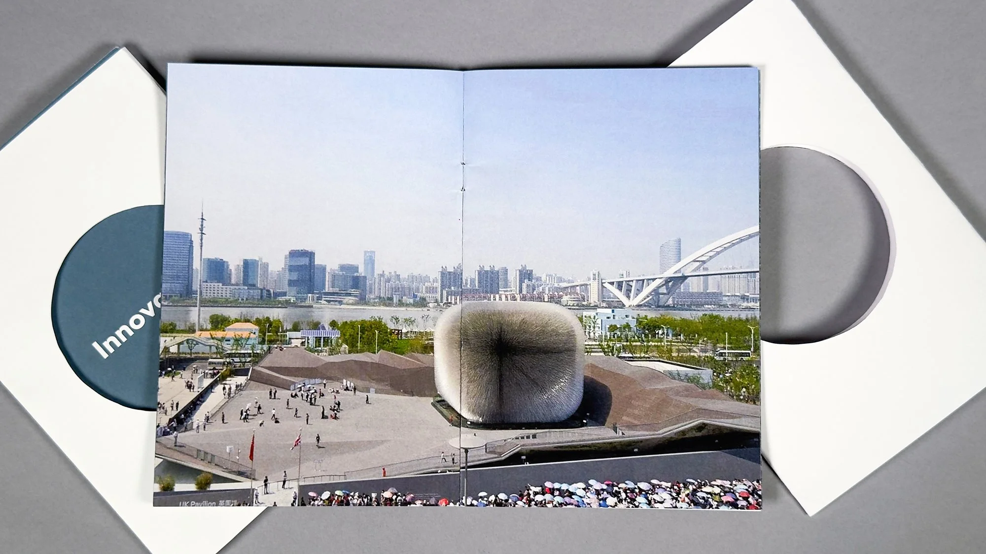 An aerial photo of the UK Pavilion in its entirety , with an urban skyline in the background and people walking in the foreground.