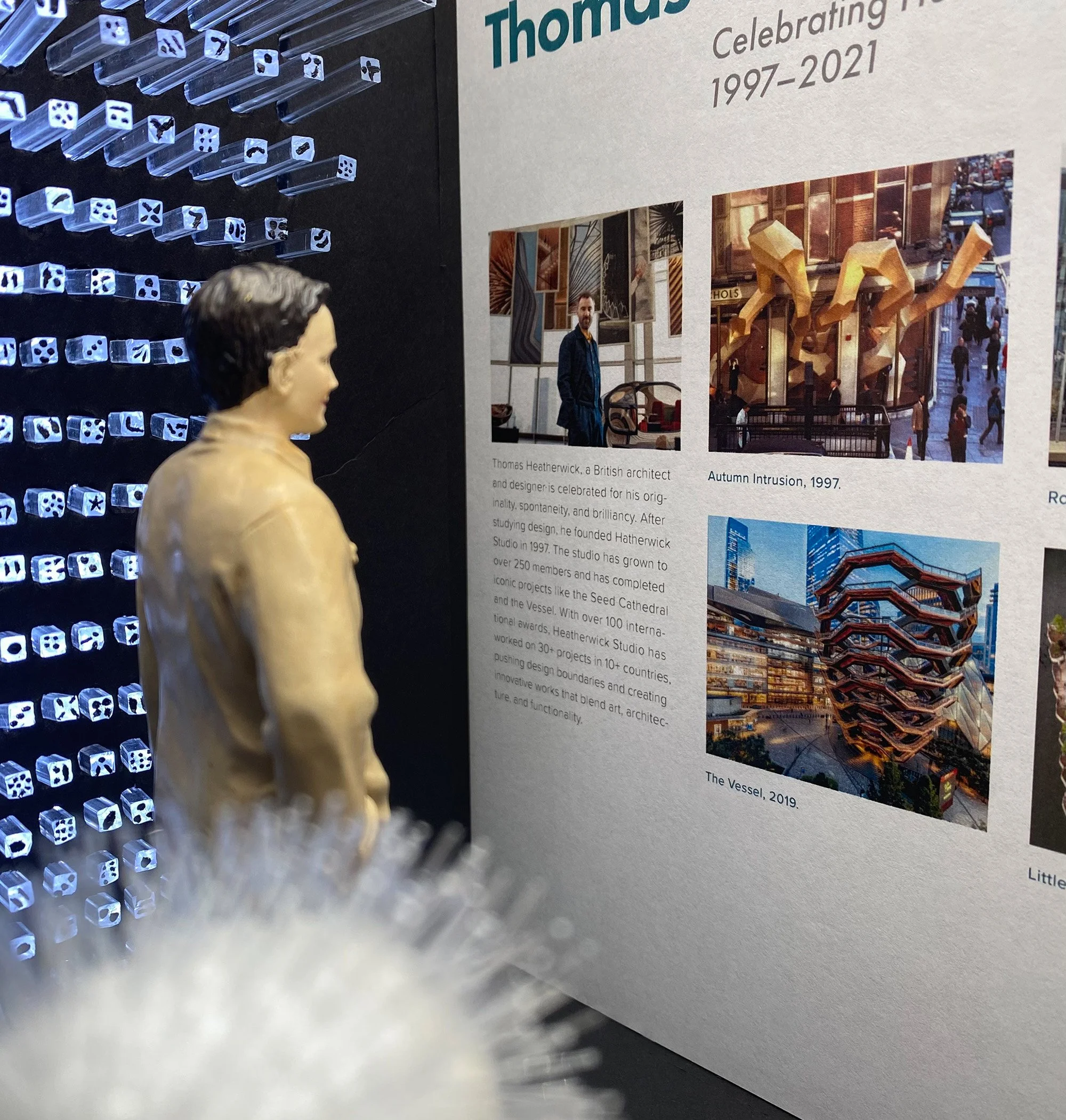 Close-up of a man standing in an art gallery looking at images about architect Thomas Heatherwick, celebrating his work from 1997 to 2021, featuring photos of public art installations.