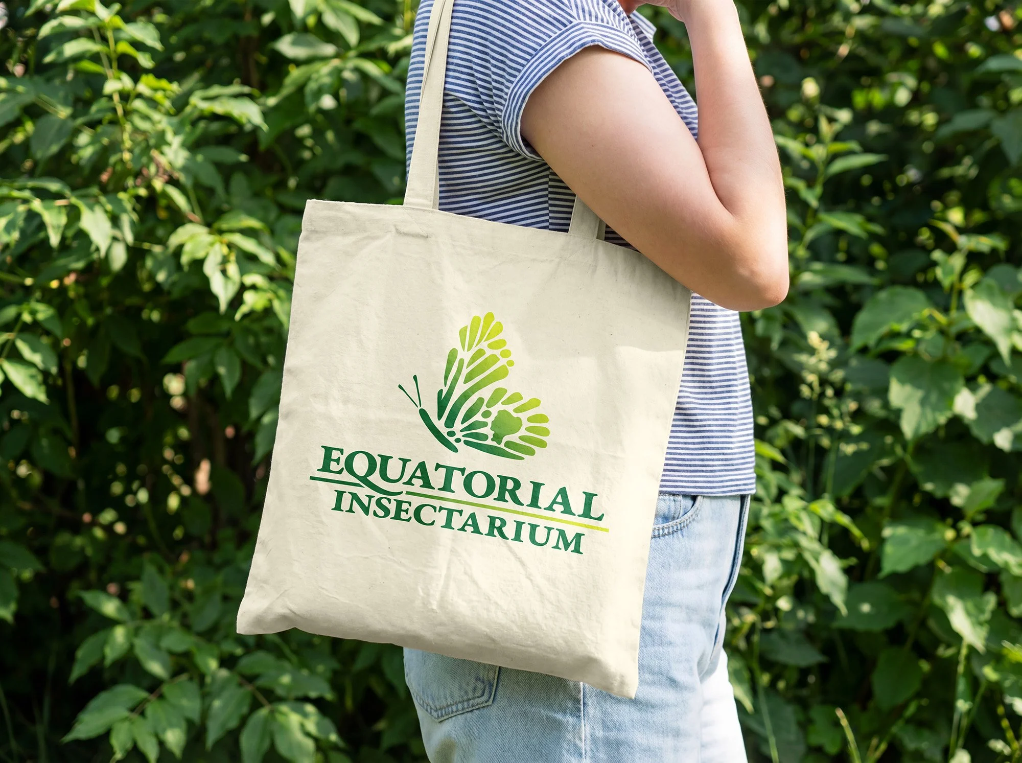 Person wearing a blue and white striped shirt, carrying a tote bag with the logo and text 'EQUATORIAL INSECTARIUM' on it, standing outdoors with a nearby shrubs in the background.