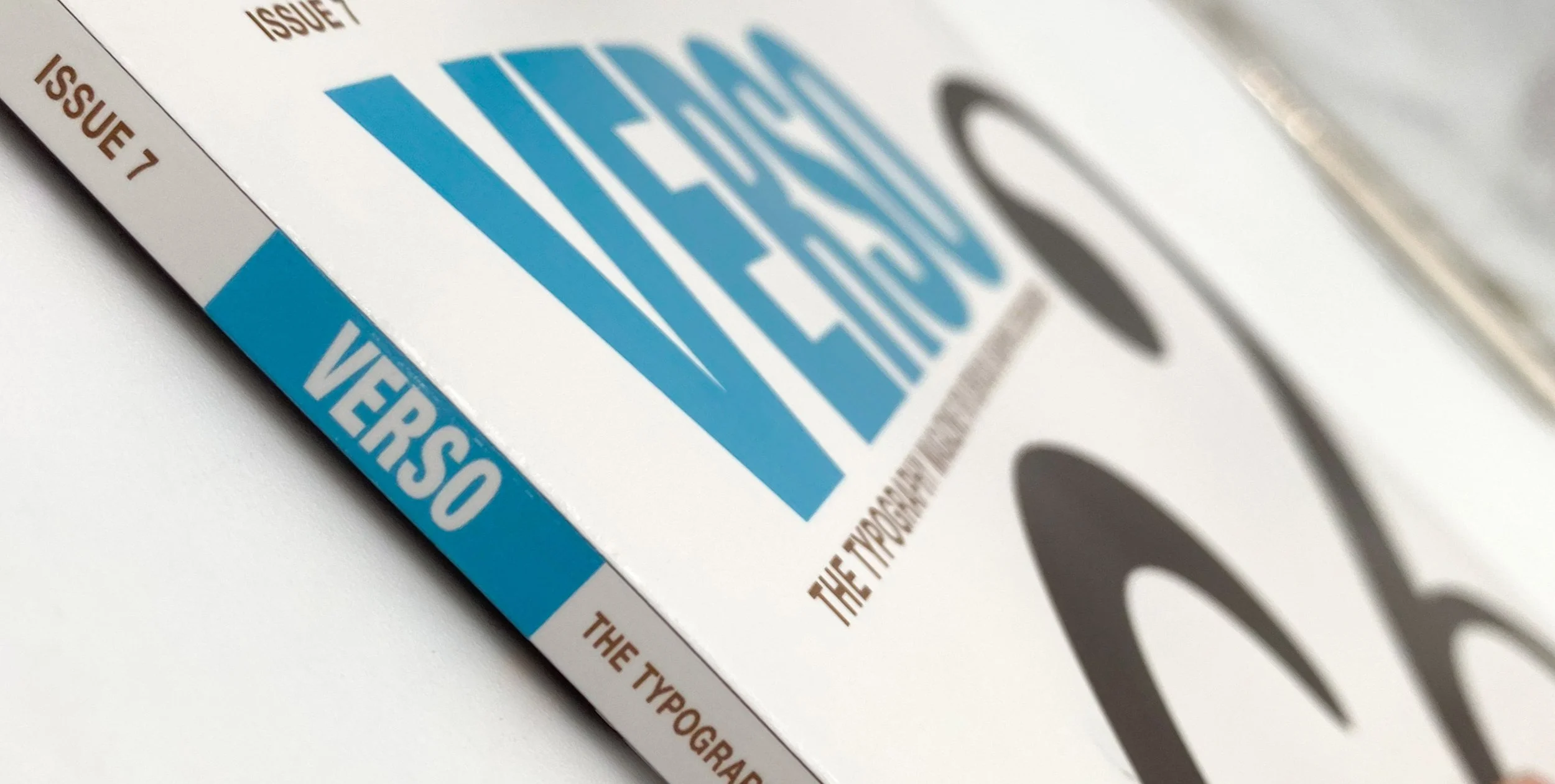 Close-up of a magazine spine that reads 'ISSUE 7', then  'VERSO' contained in a teal blue square while the cover shown is blurry but also reads "VERSO" in large teal blue letters and shows an interesting dark brown shape under the title.