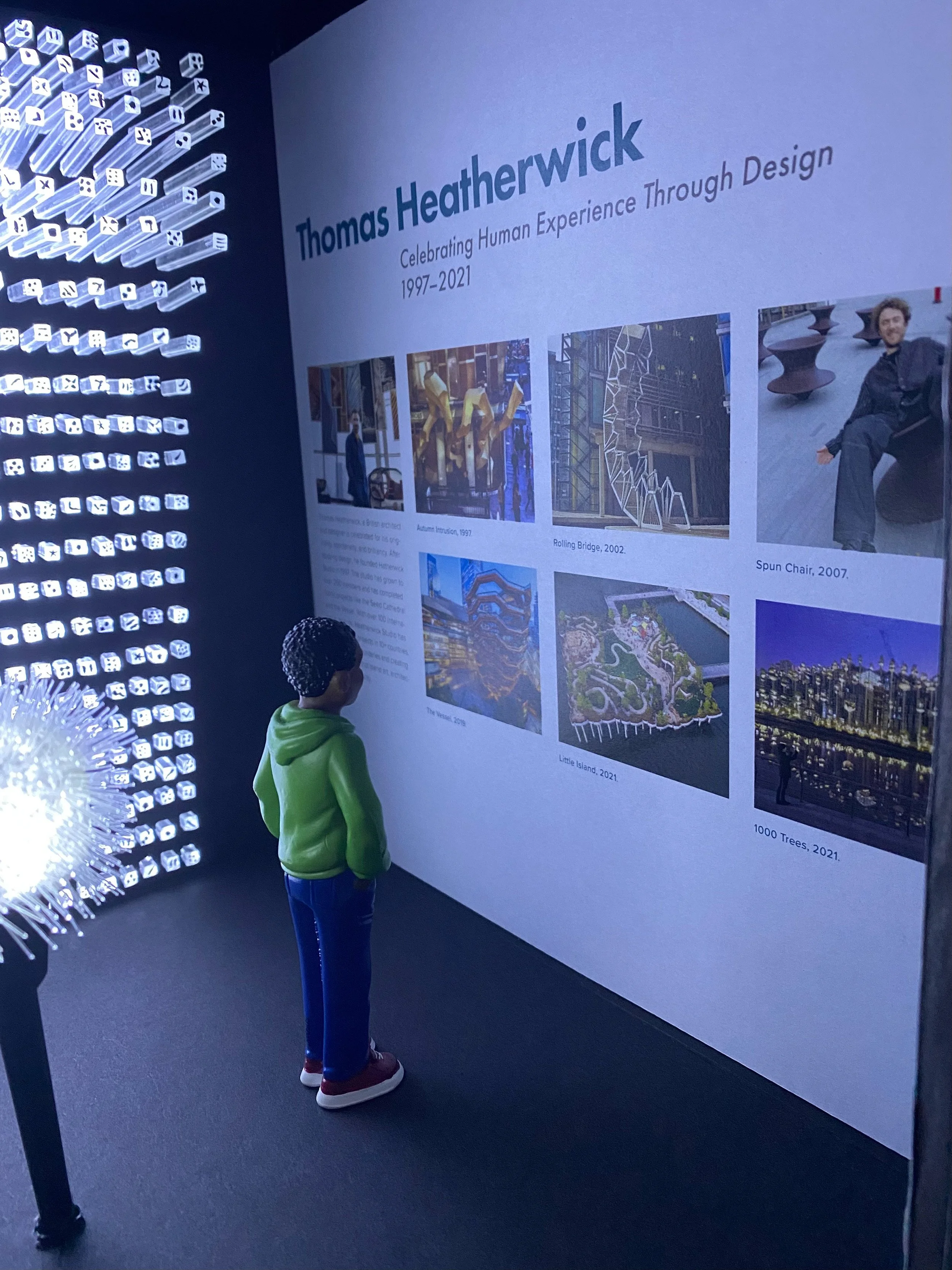 A museum exhibit dedicated to Thomas Heatherwick, displaying text and pictures of his projects from 1997 to 2021, with a child observing and reading it.