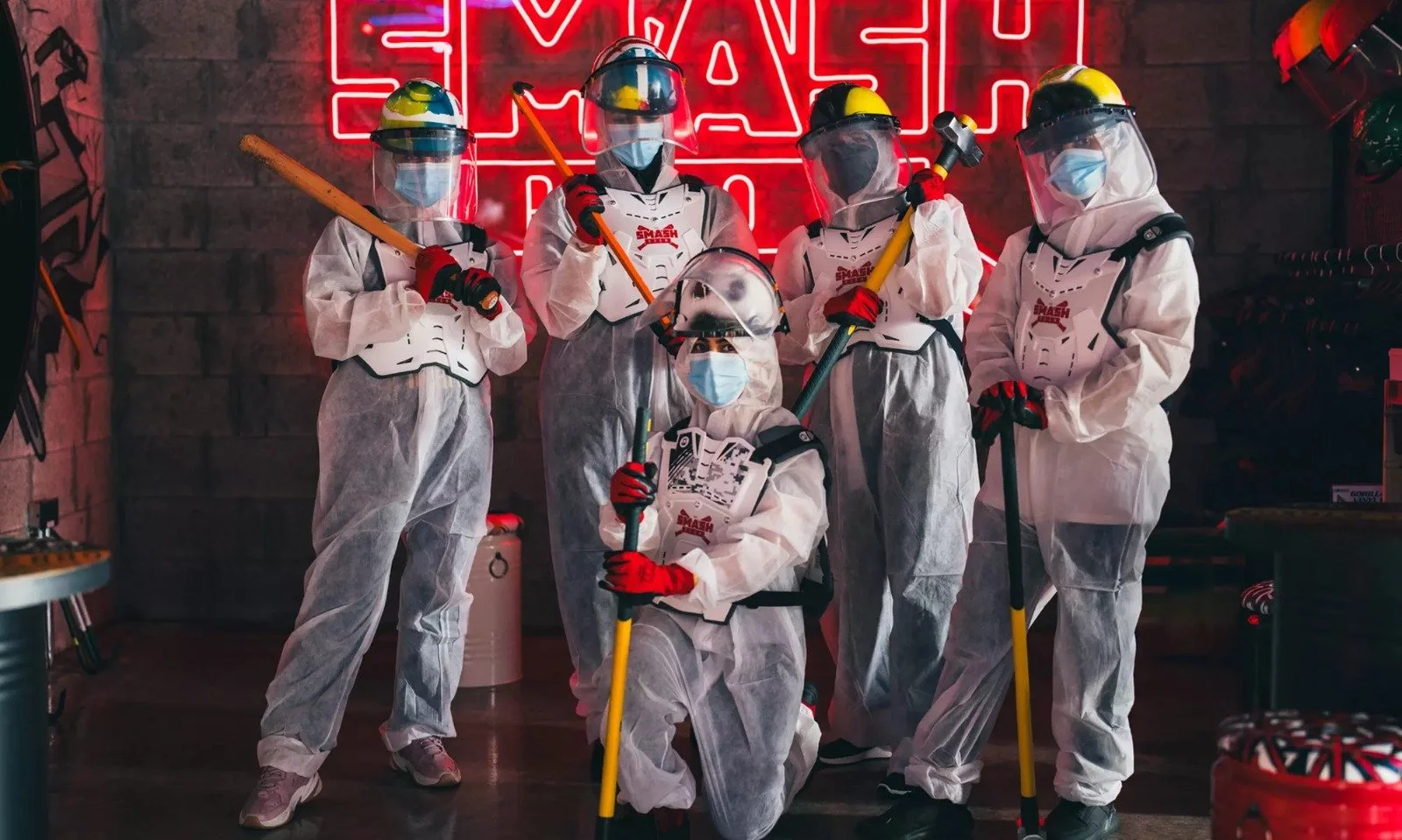 People dressed in white hazmat suits with face masks, helmets, and face shields, holding cleaning tools in a room with a red neon sign in the background.