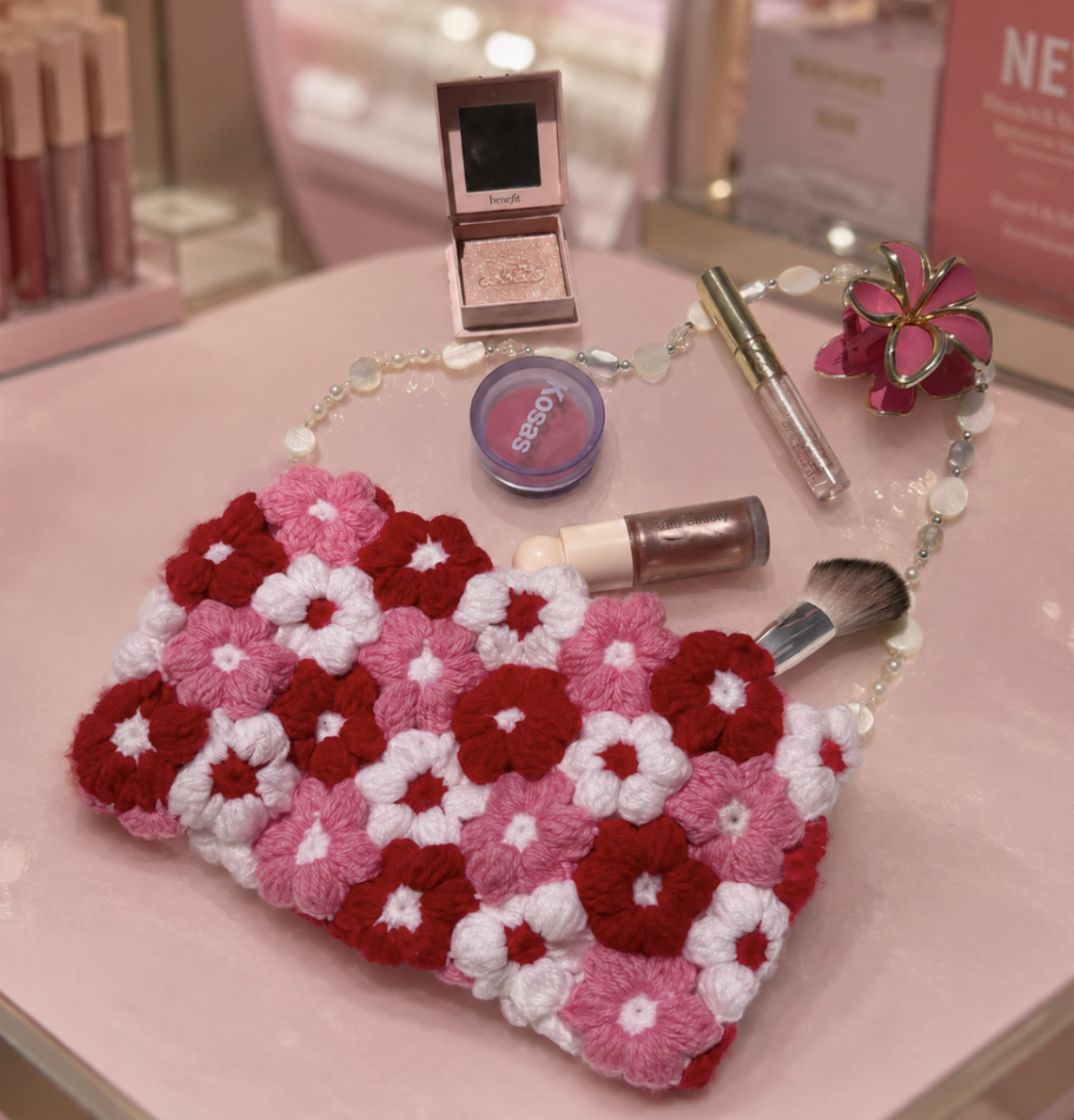A pink heart-shaped bag made of yarn flowers in shades of pink, red, and white, surrounded by makeup and beauty accessories including a Benefit eyeshadow palette, a small jar of lip gloss, a blush compact, makeup brushes, and a decorative pink flower-shaped container, all arranged on a pink surface.