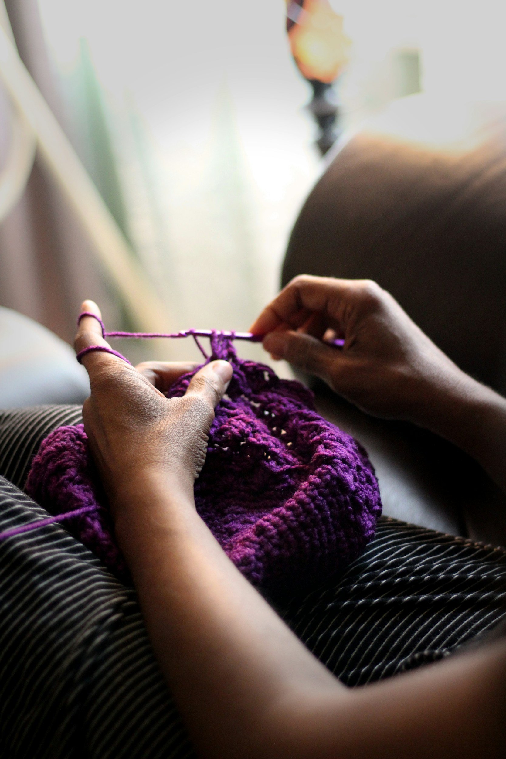 Close-up of hands knitting purple yarn, with a cozy purple knitted item in the process. Someone is sitting indoors, wearing striped pants, with a blurred background.