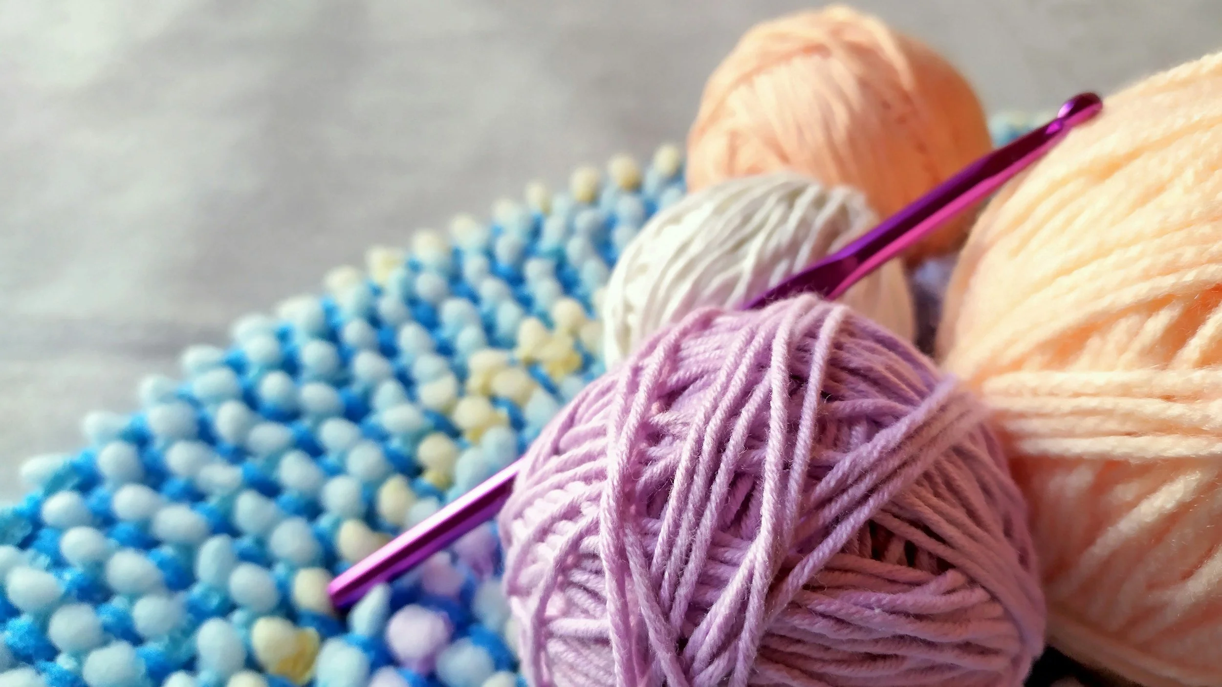 Close-up of various balls of yarn in purple, peach, white, and pink, with a knitting needle inserted into the pink yarn.