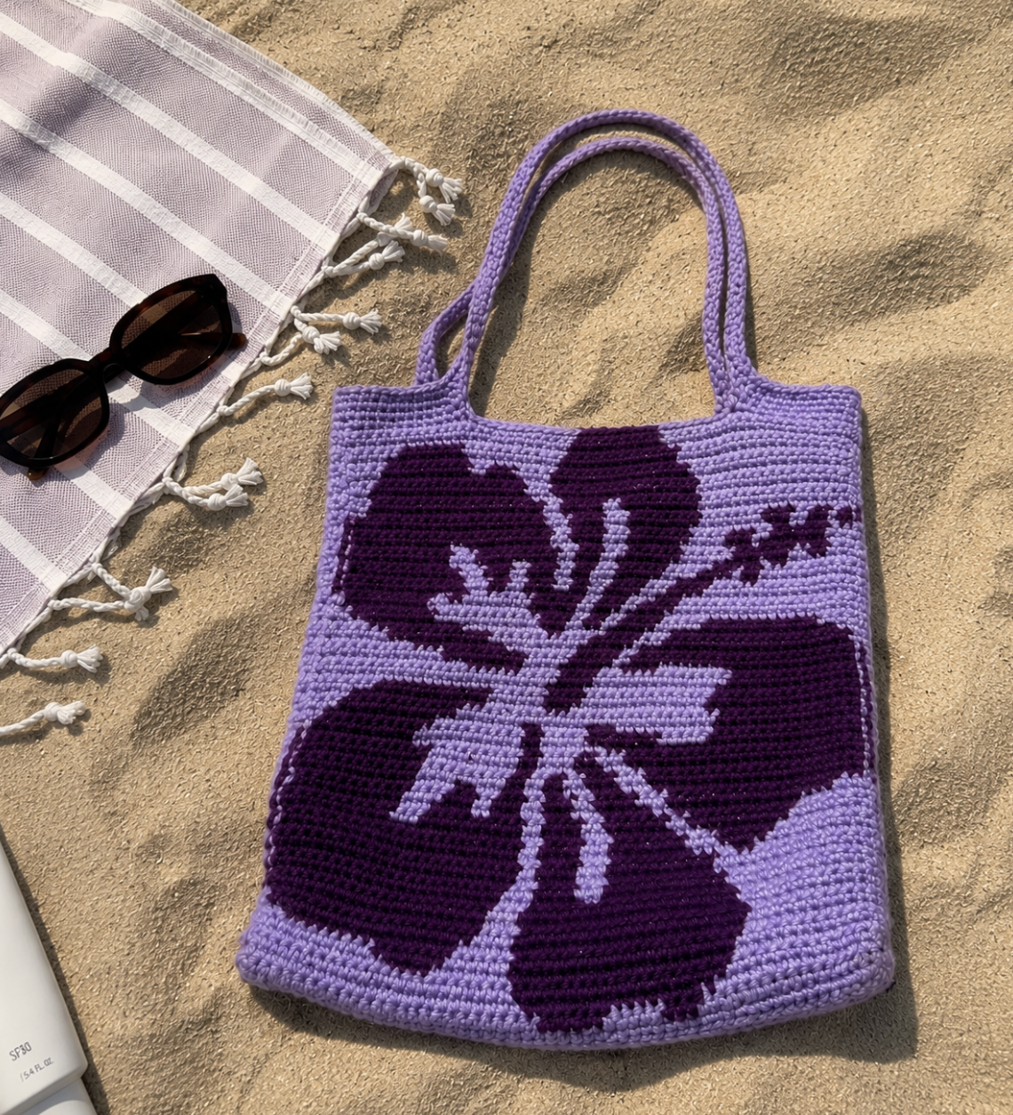 A purple and violet floral crocheted tote bag on sandy ground near a white beach towel with tassels and a pair of sunglasses.