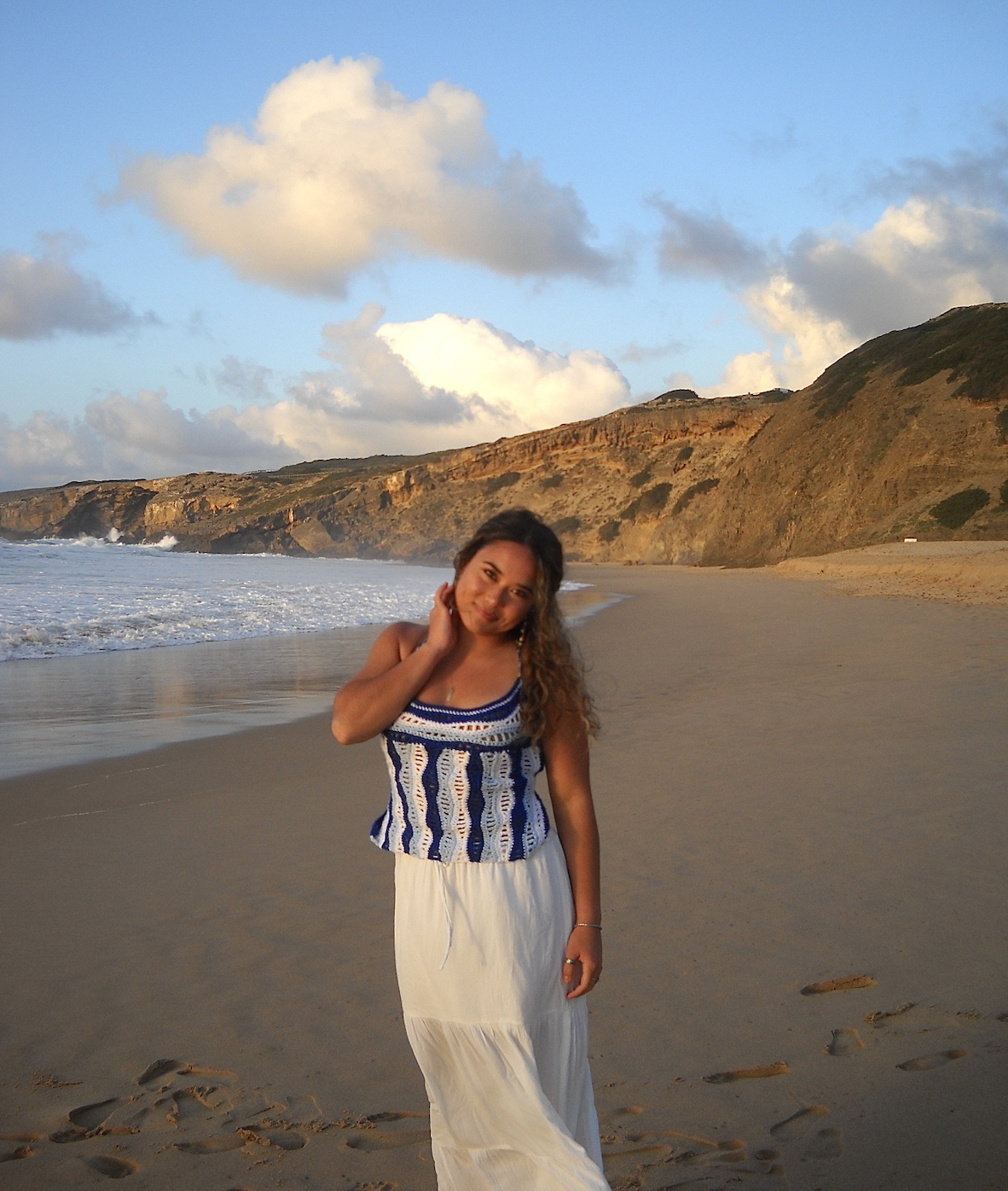 A woman with long, curly hair standing on a sandy beach during sunset with cliffs and clouds in the background.