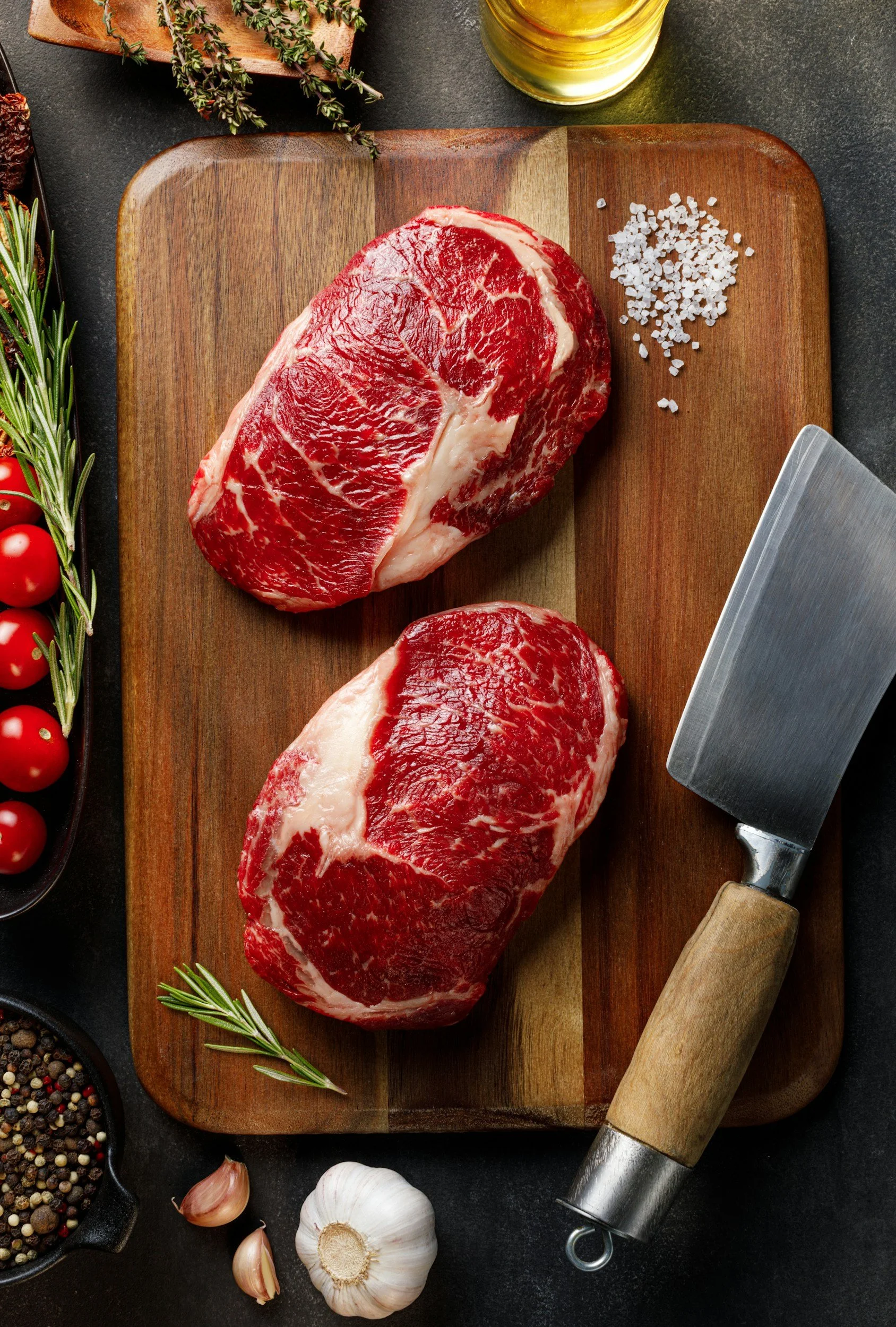 Two raw beef steaks on a wooden cutting board, surrounded by garlic, cherry tomatoes, fresh herbs, salt, pepper, and a meat tenderizer on a dark surface.